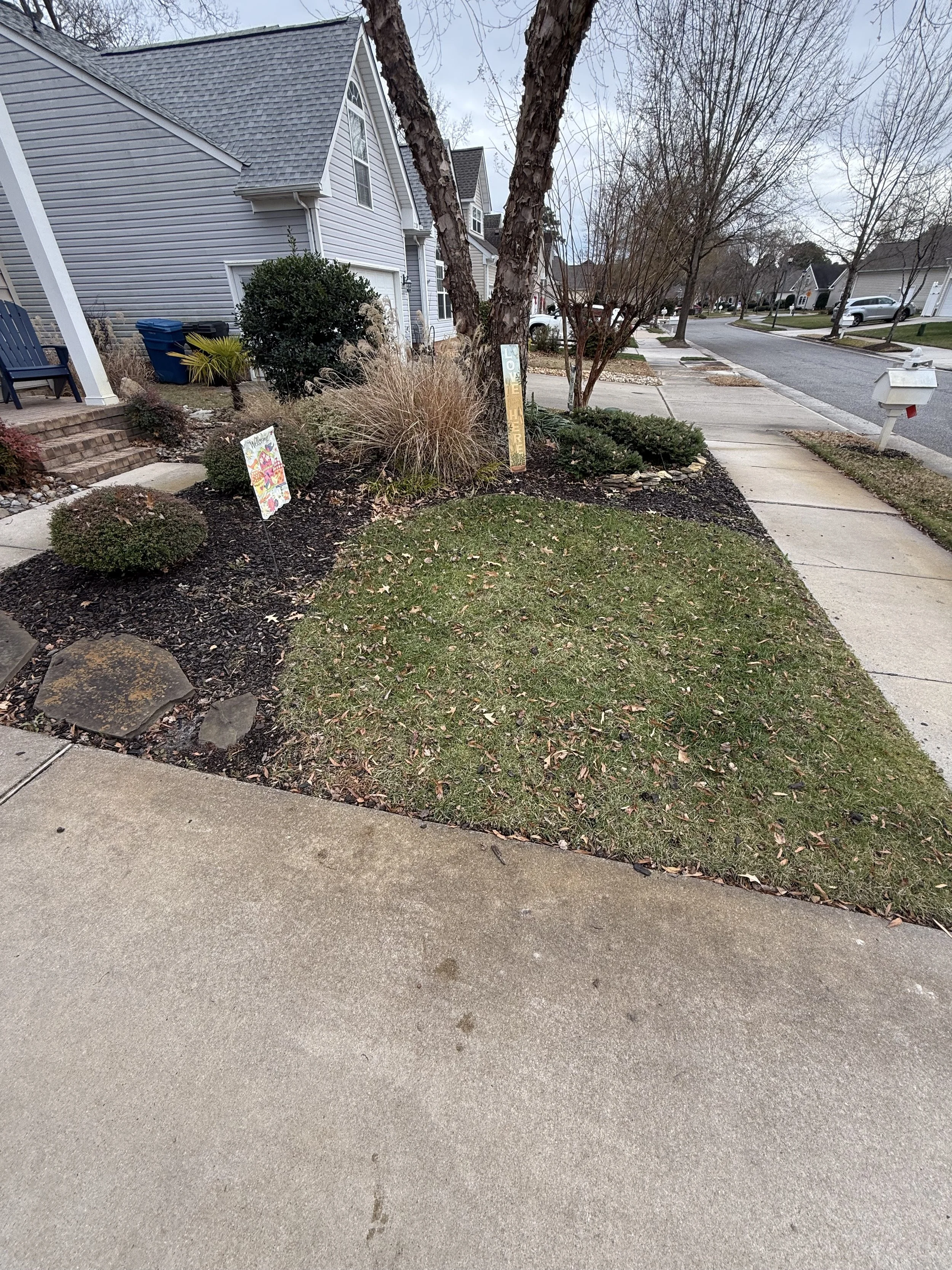 A front yard with a small grass patch surrounded by mulch and a concrete sidewalk. There are two signs in the yard, one painted and one wooden with the words 'Love' and 'Here' visible. There are trees with bare branches, bushes, and part of a house w