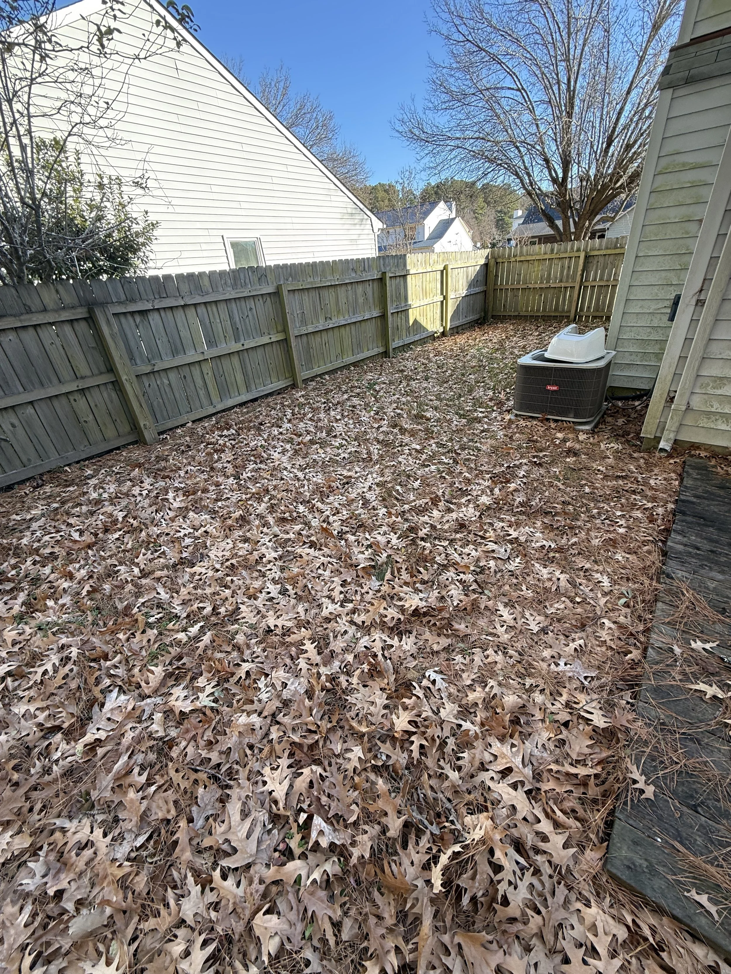 Backyard with fallen leaves on the ground, surrounded by a wooden fence, with houses and trees in the background, and an HVAC unit near the house.