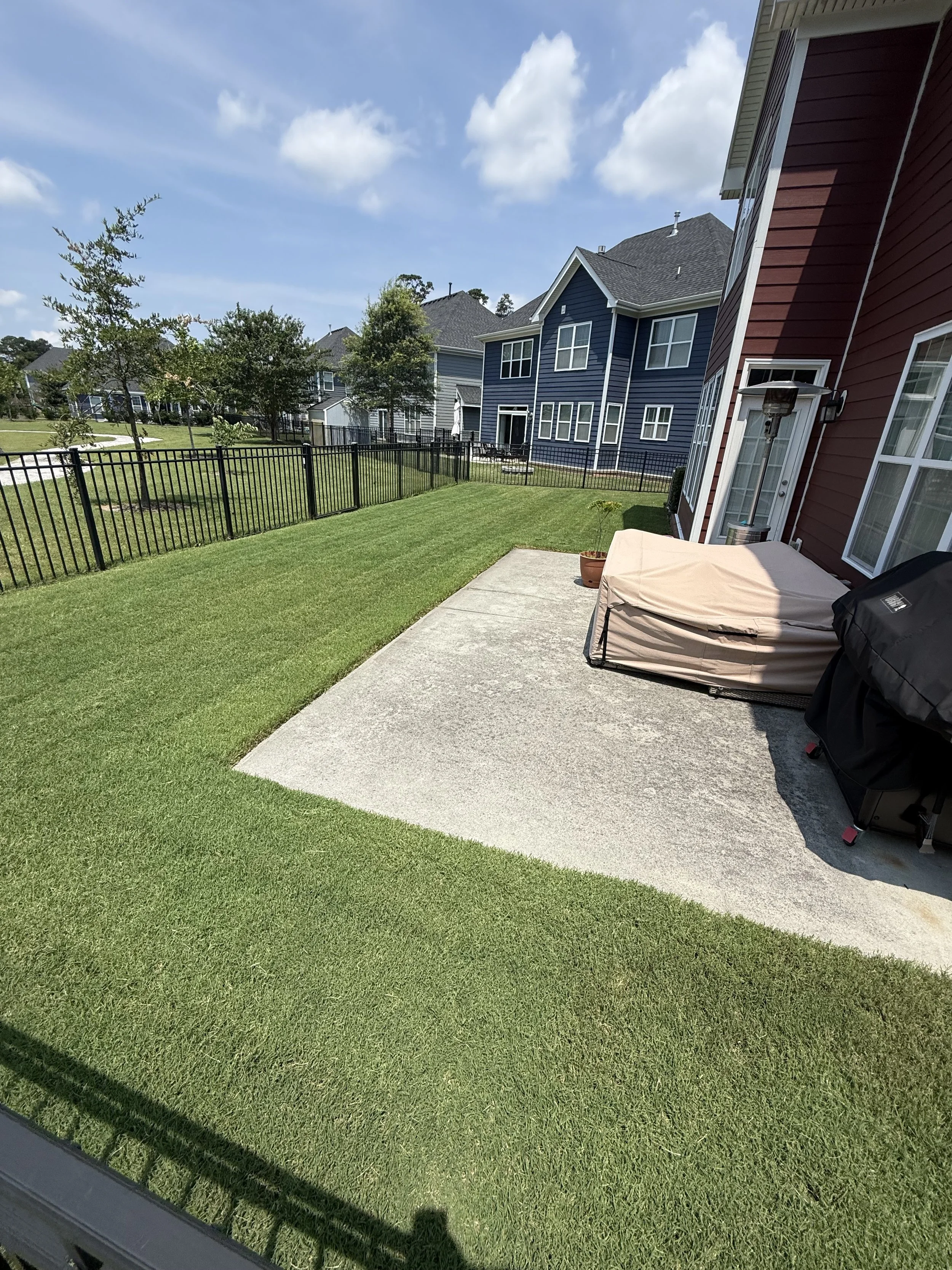 A backyard patio with a concrete slab, a covered grill, potted plant, and patio heater, fenced yard, green grass, neighboring houses, blue sky with clouds.