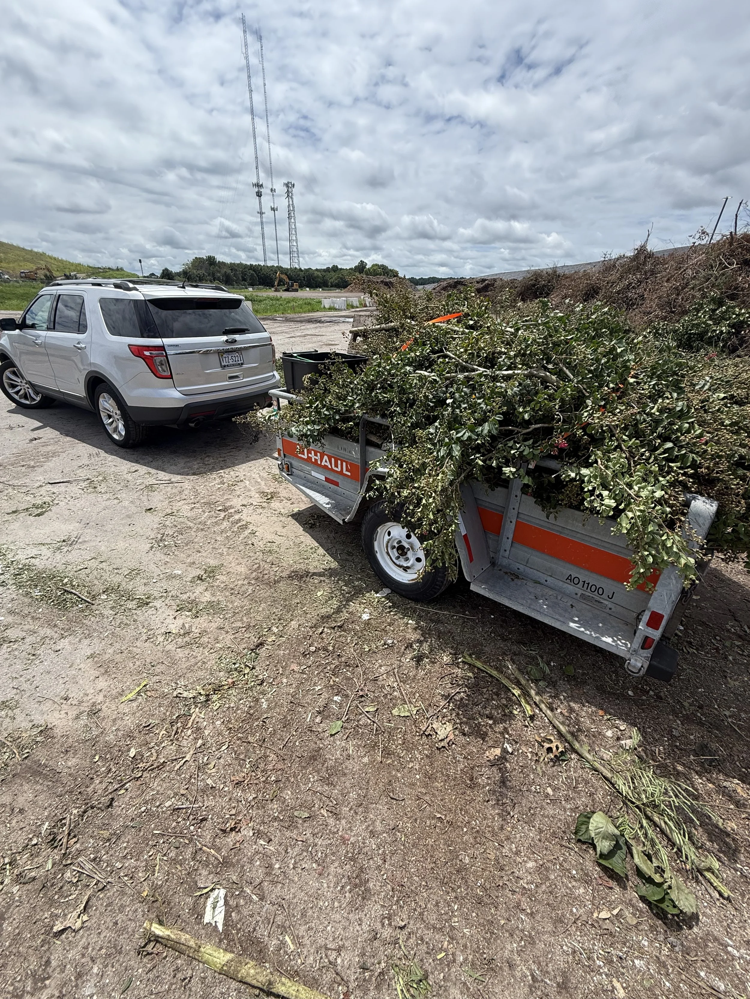 A silver SUV parked on dirt with a U-Haul trailer attached, filled with branches and leaves. There are utility poles and a cloudy sky in the background.