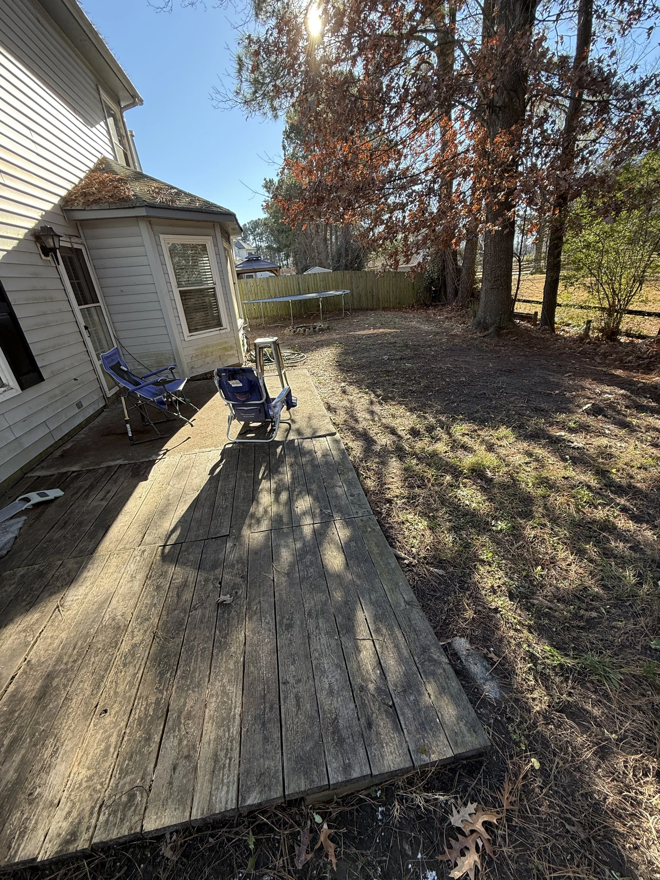 Backyard patio with wooden deck, chairs, a trampoline, and a large tree, with a house on the left and a wooden fence in the background under a sunny sky.