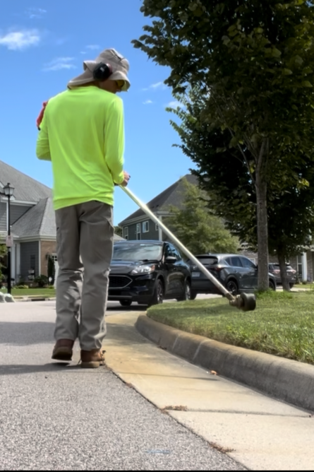 A man wearing a neon yellow long-sleeve shirt, gray pants, a wide-brimmed hat, and headphones is trimming grass along a sidewalk with a weed trimmer on a sunny day. Houses and parked cars are visible in the background.