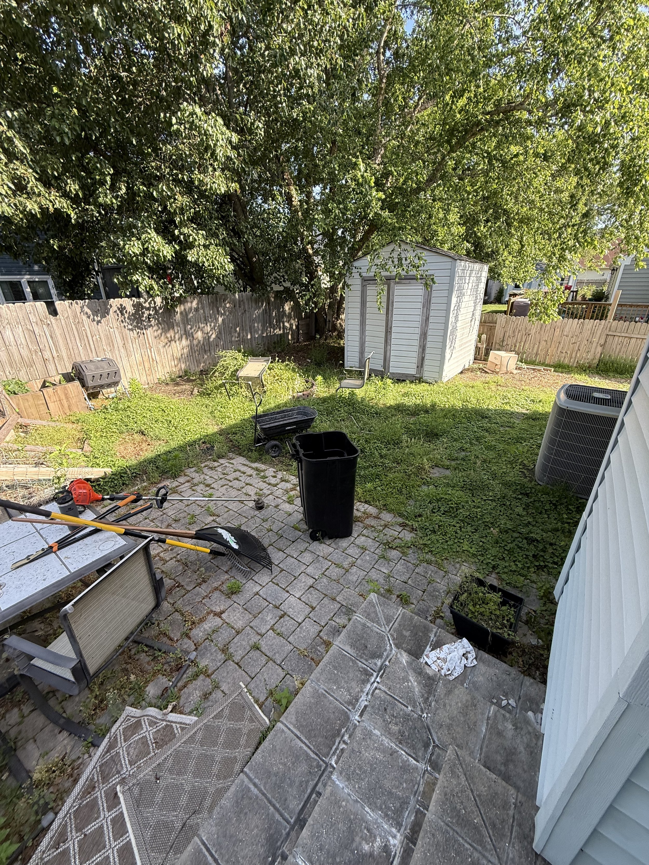 Backyard with paving stones, lawn, garden shed, tree, fence, and outdoor items including trash bin, garden rake, and a patio furniture set.