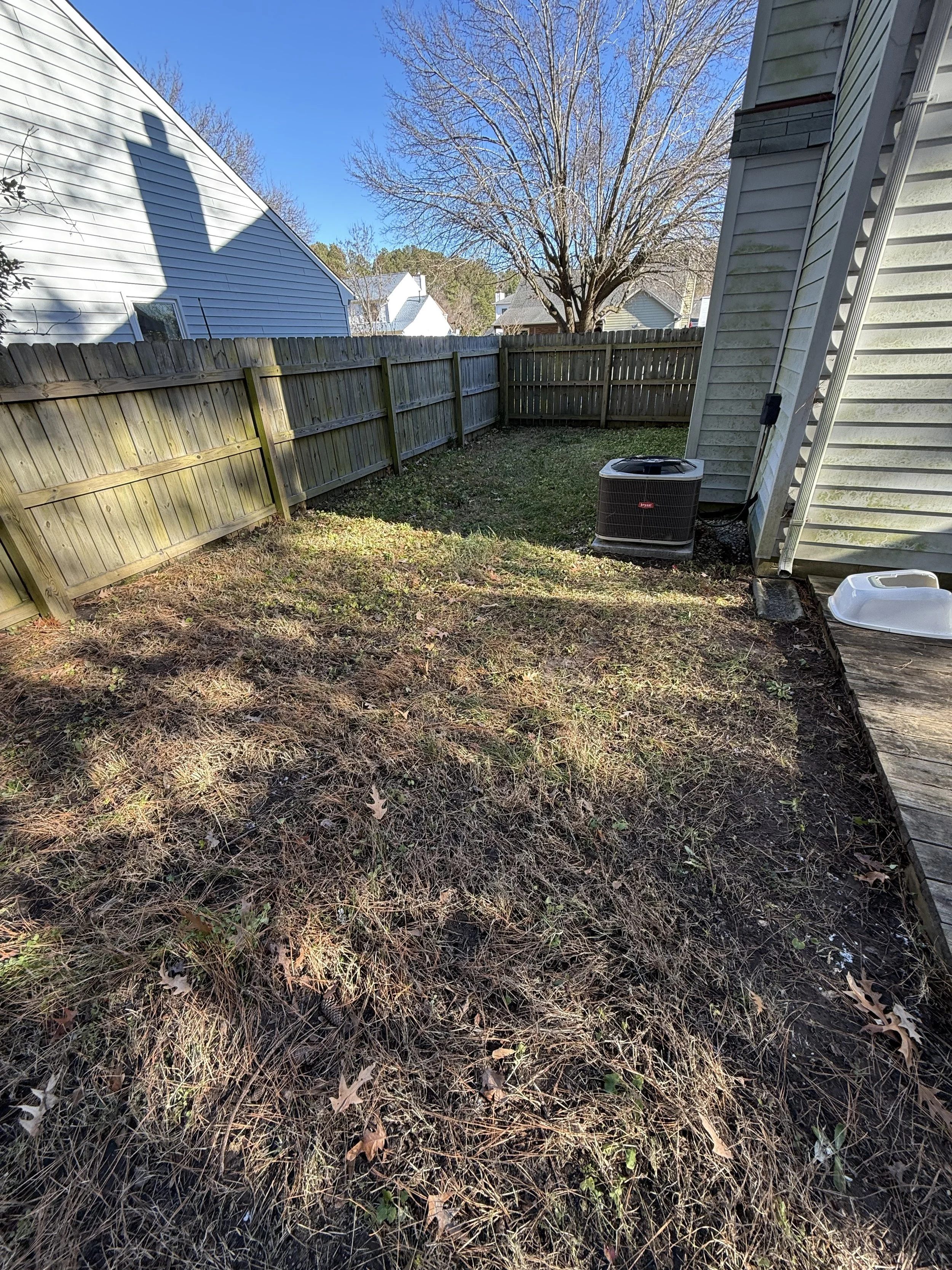 Backyard with a wooden fence on three sides, a small patch of grass, and a deck on the right. An air conditioning unit is positioned against the house, with a white feed tray on the deck nearby. A large tree with no leaves is visible in the backgroun