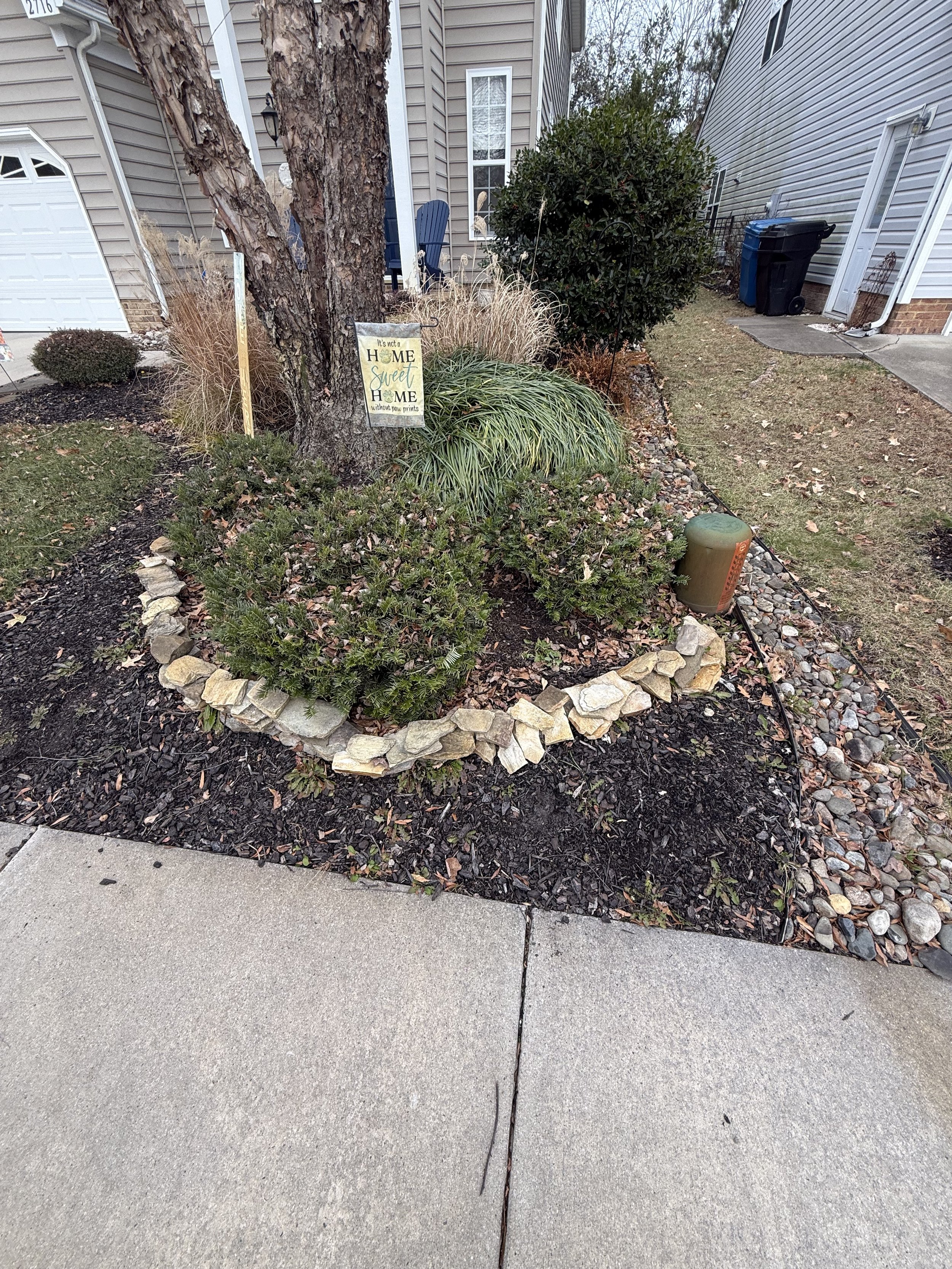 A small landscaped garden bed with a tree, bushes, ornamental grasses, decorative rock border. Located near a sidewalk and adjacent to a residential house.