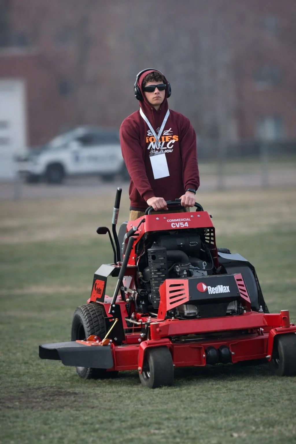 A young person wearing sunglasses, a hoodie, and a maroon sweatshirt operating a red riding lawn mower outdoors on grass. They have a lanyard with a badge around their neck and are wearing a headset.