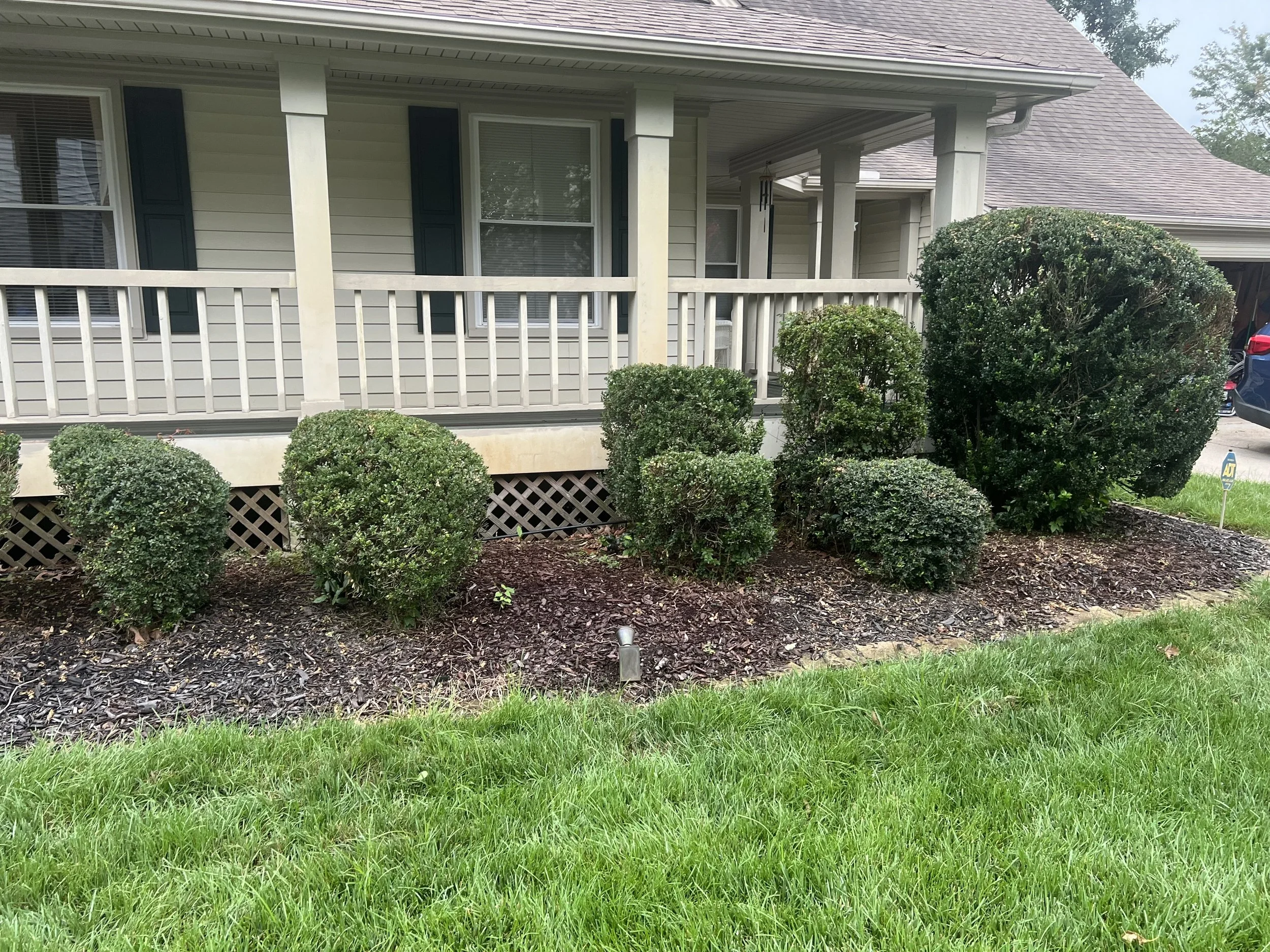 Front yard with well-maintained bushes and grass in front of a house with a porch and white railing.