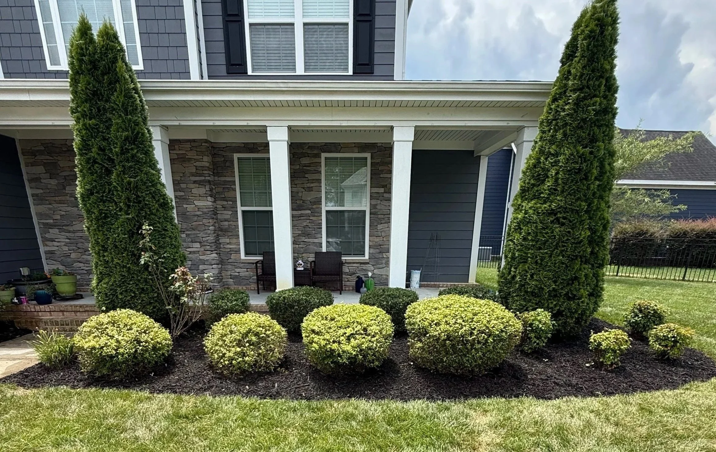 Front porch of a house with stone and navy blue siding, two large tall evergreen trees on either side, and well-maintained bushes and grass in front.
