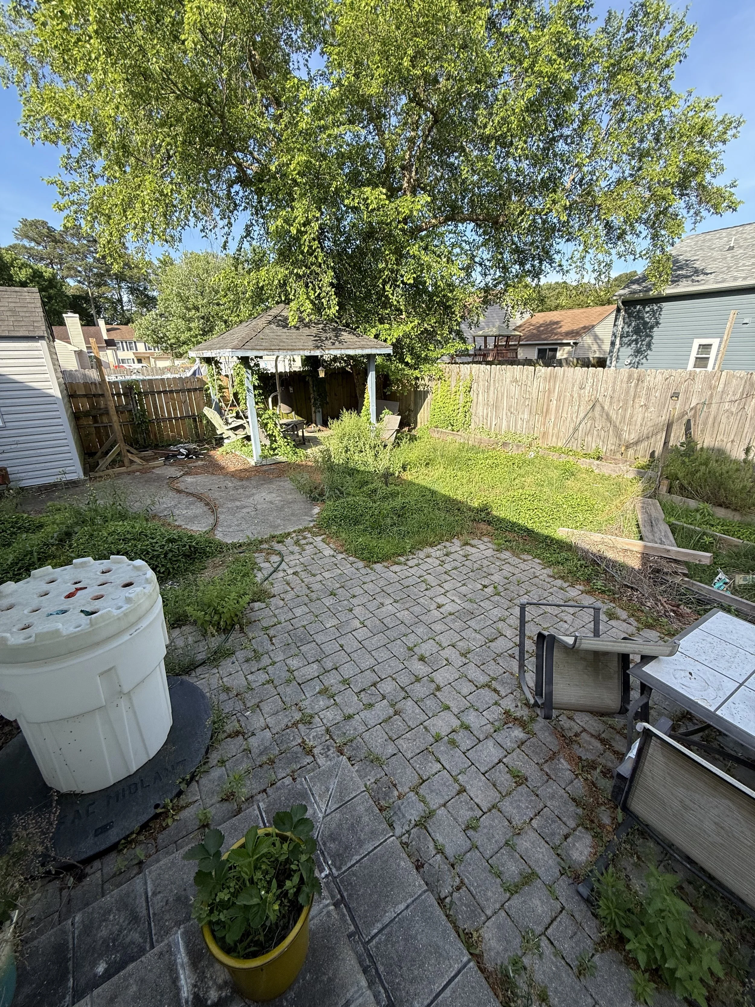 A backyard with a large tree, a brick patio, a covered gazebo, outdoor furniture, a potted plant, and a row of houses in the background.