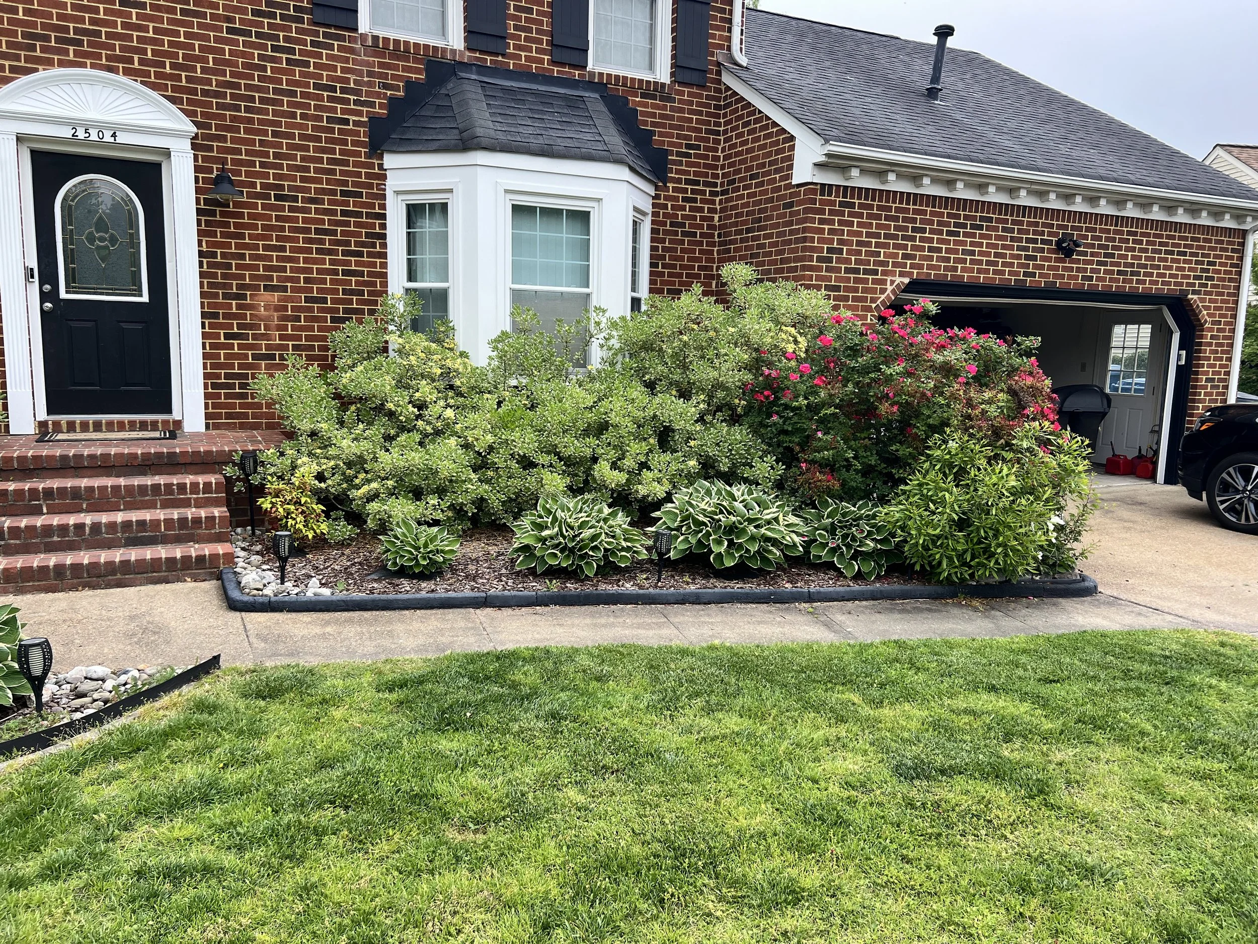 A brick house with front steps, black front door, and a garden bed with green bushes, pink and white flowers, hostas, and decorative lighting along the sidewalk.