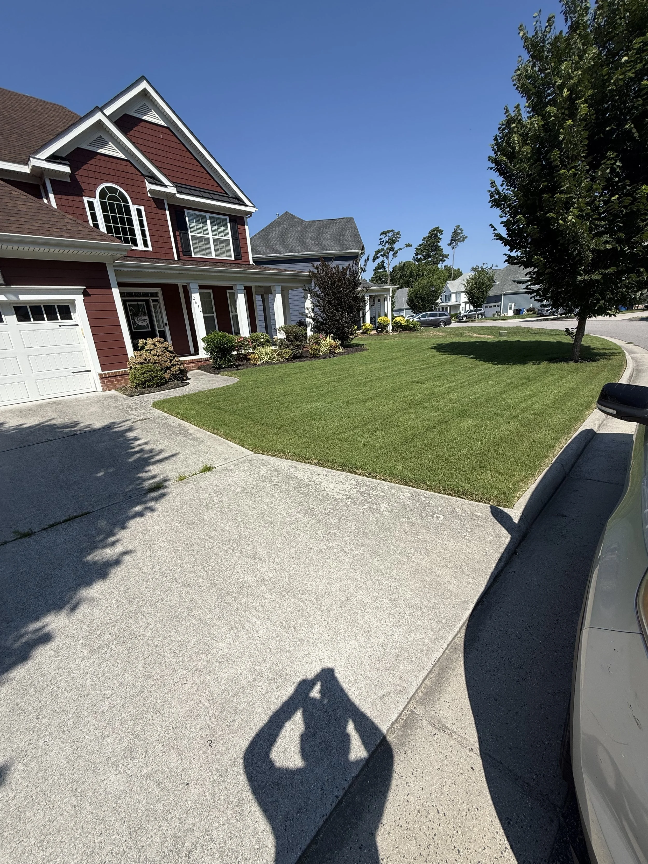 A suburban house with red siding and white trim, a well-manicured front lawn, and a driveway with a shadow of the photographer taking the picture on a sunny day.