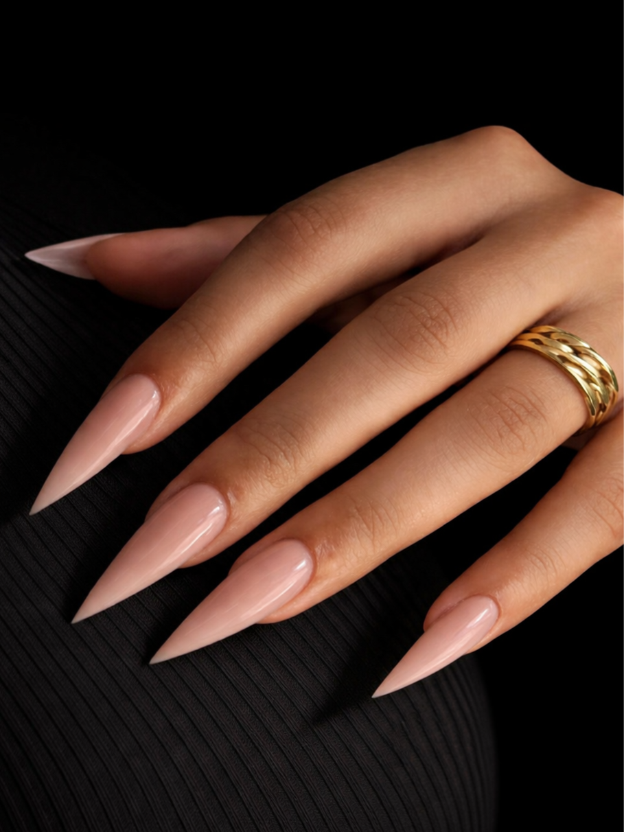 Close-up of a woman's hand with long, pointed, nude-colored acrylic nails and a gold ring, resting on a black textured surface.