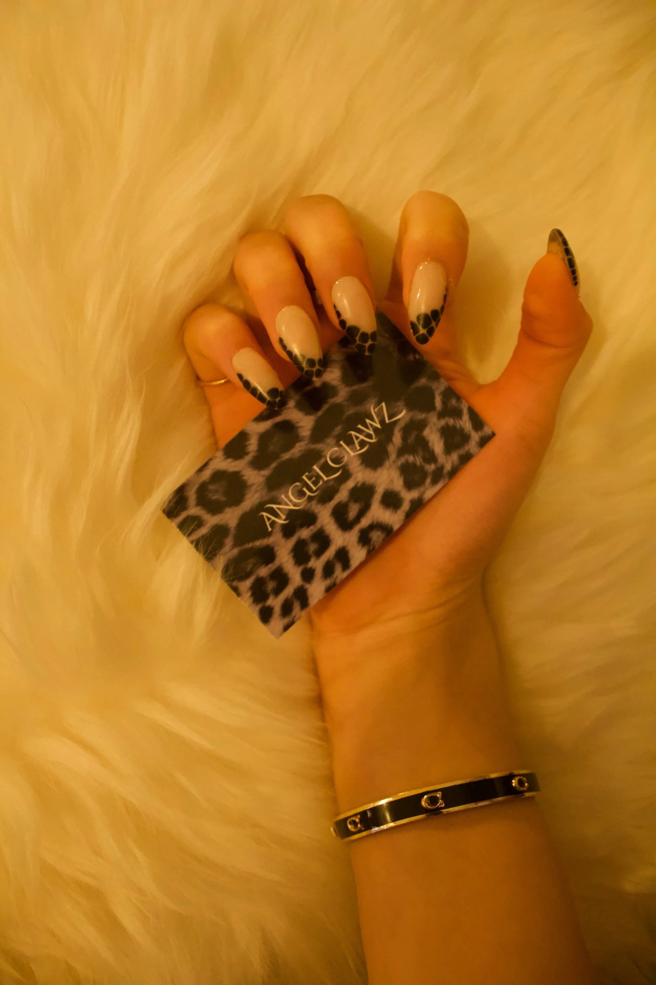 A hand with leopard print nails holding a business card with a leopard print background and the words 'Angel Clawz' written on it, resting on a fluffy white fur surface, and wearing a black and gold bracelet.