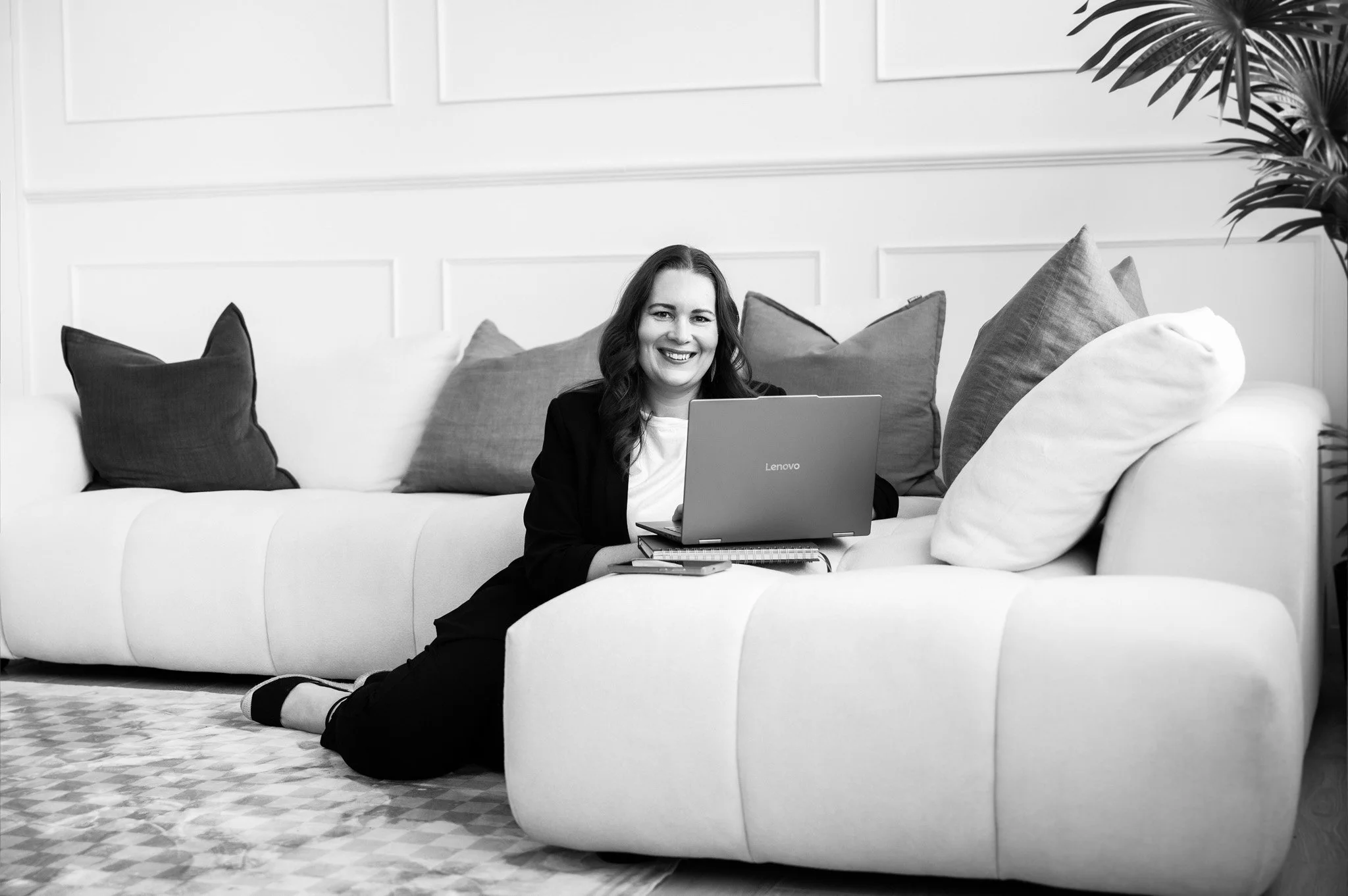 Black and white image of Tracey sitting on the floor next to a couch, working with her laptop and smiling