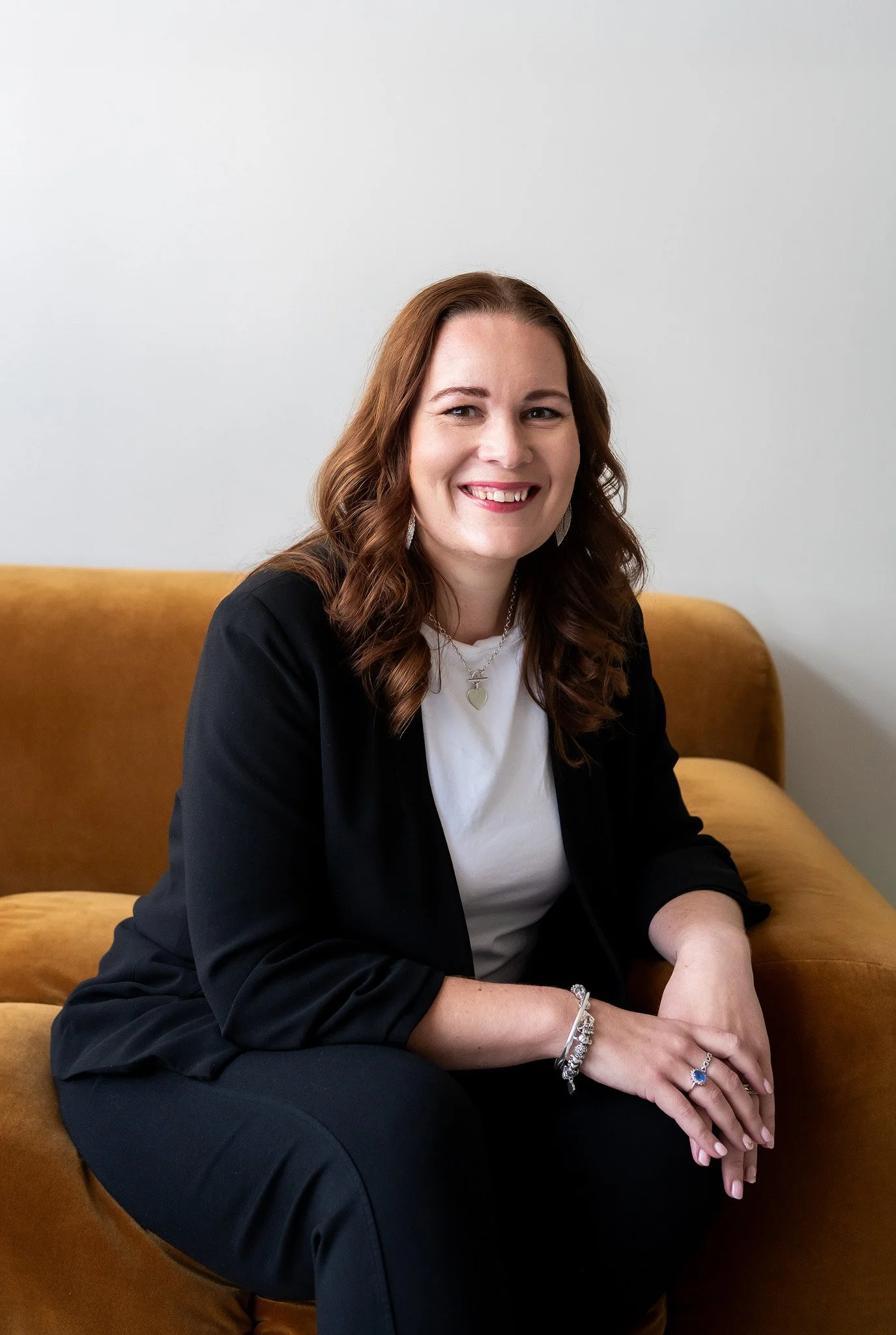 Tracey sitting on a couch wearing a black blazer, white t-shirt and smiling