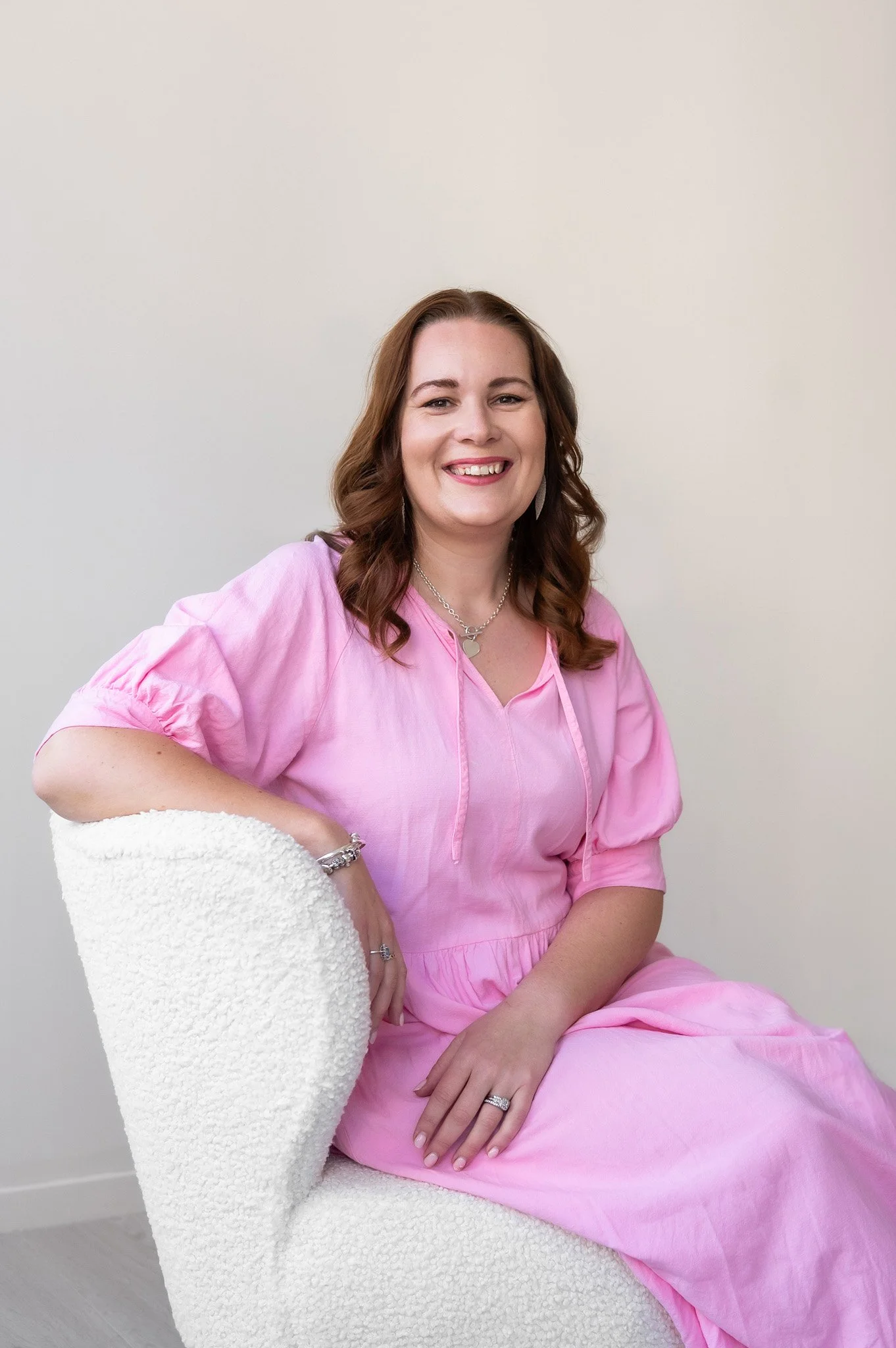 Tracey Lock sitting on a cream armchair, wearing a pink dress and smiling