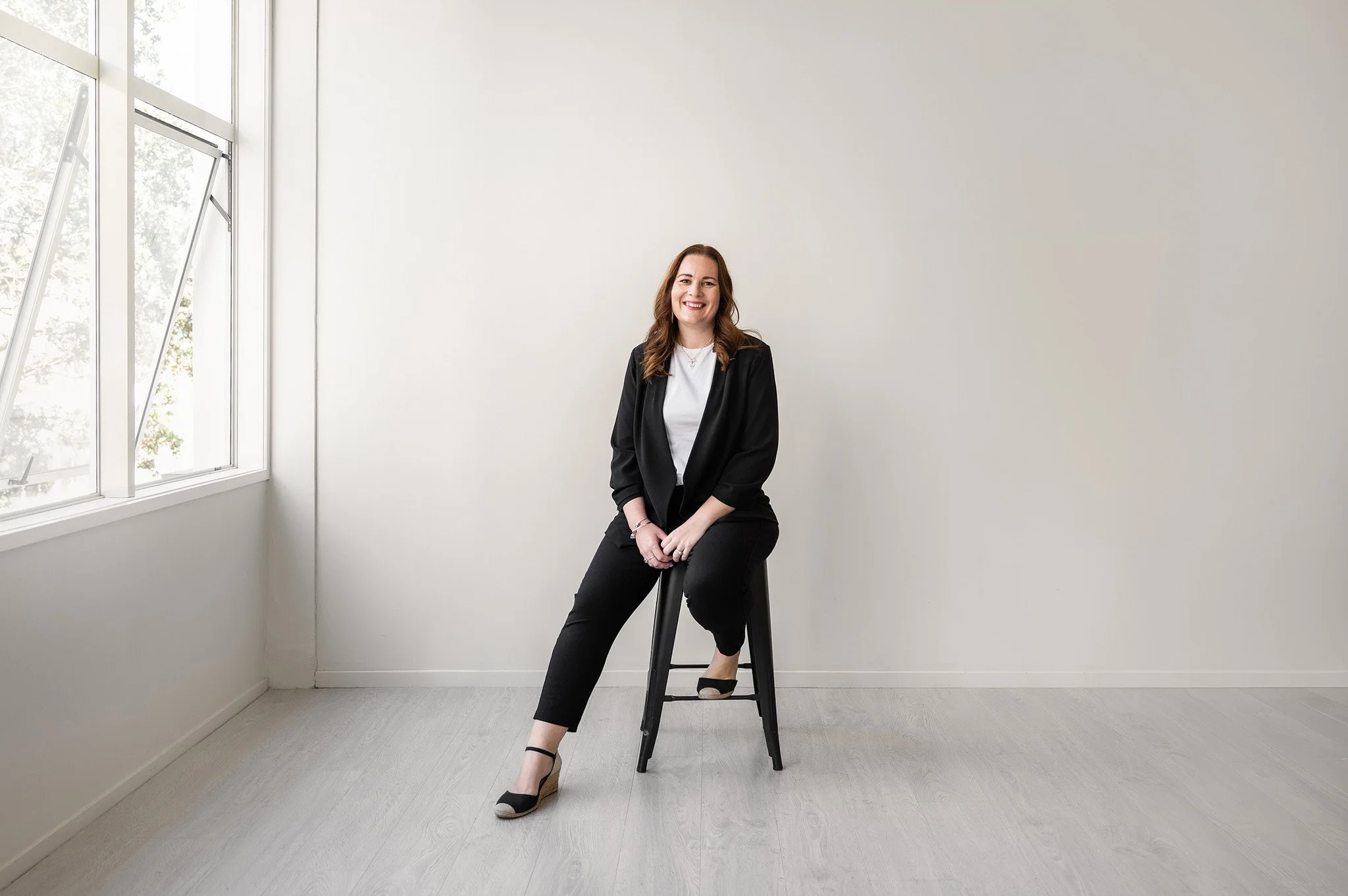 Tracey sitting on a black stool in a white studio setting, wear a black blazer, pants and white t-shirt