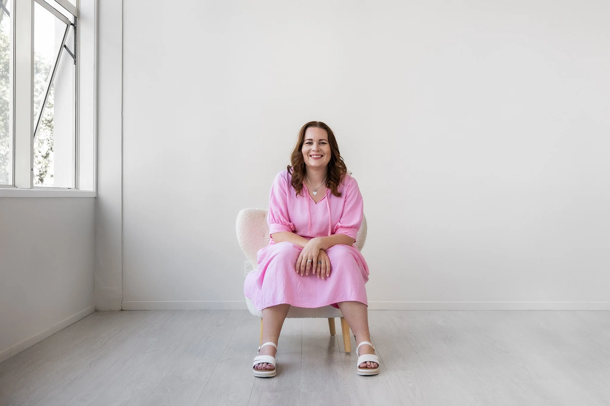 Tracey sitting on a chair facing the camera, wearing a pink dress in a white studio setting