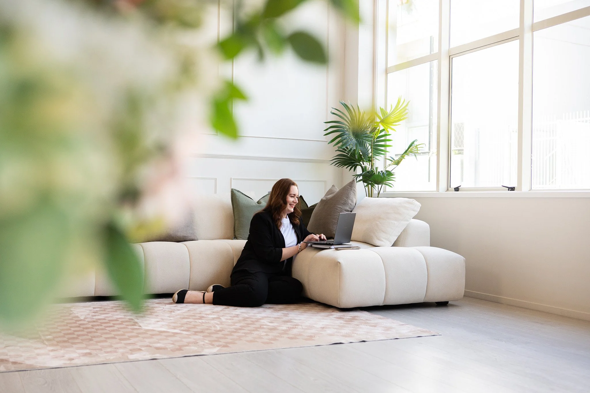 Tracey sitting on the ground working with her laptop on the couch, with greenery in the foreground of the shot