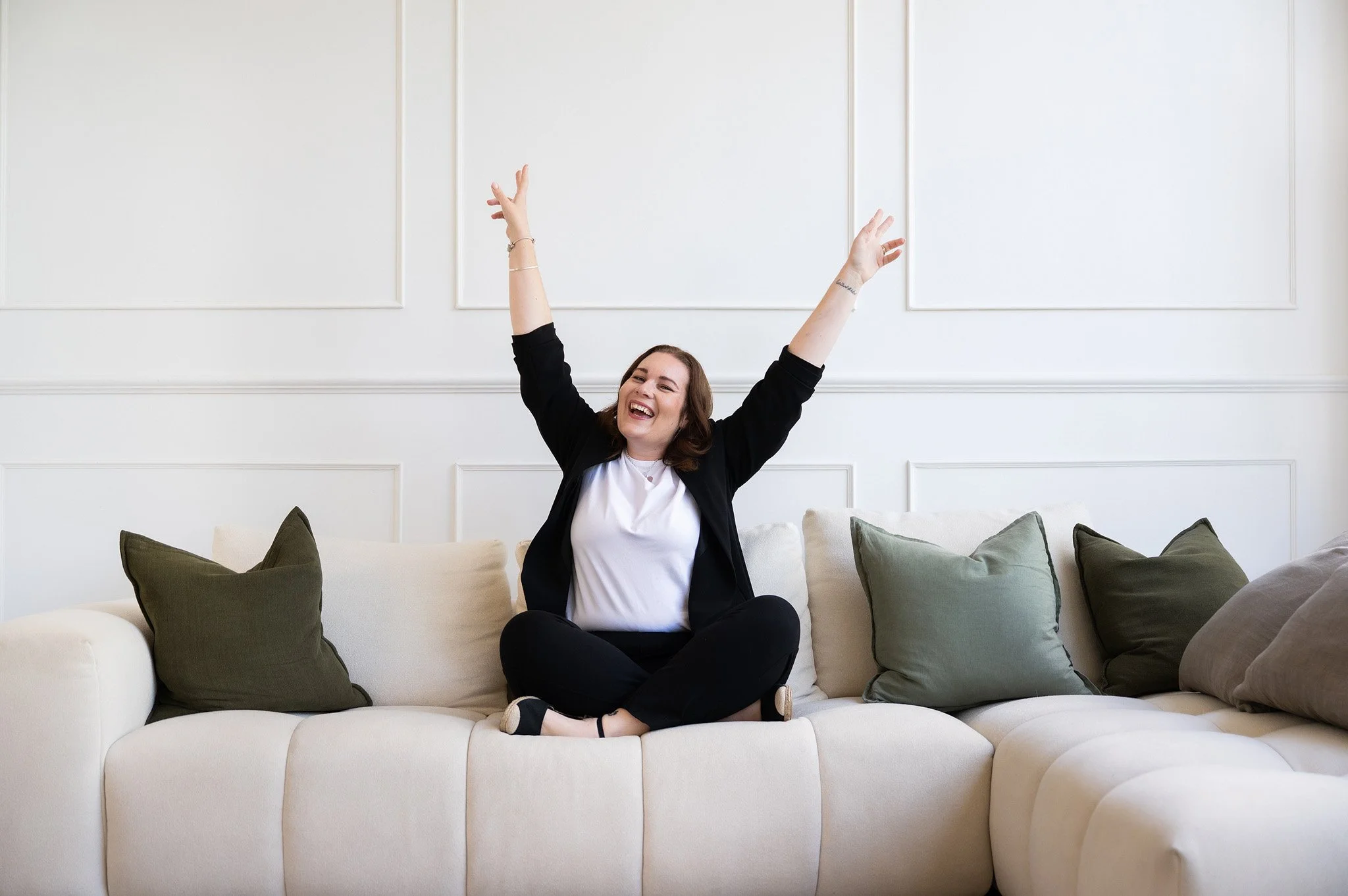 Tracey sitting on the couch cross-legged with her arms in the air and a smile on her face
