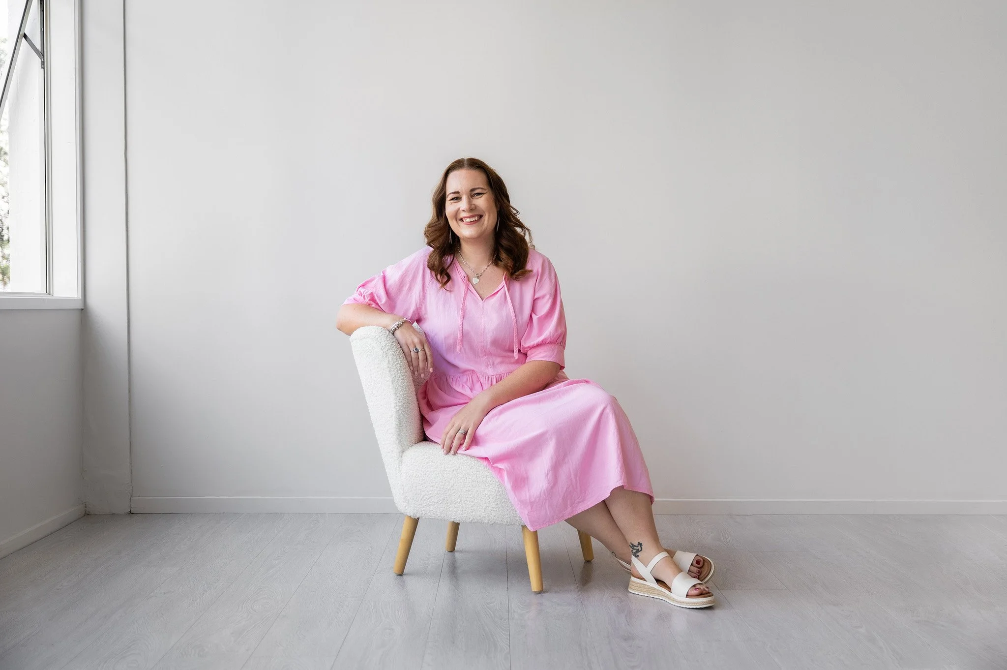 Tracey sitting on a chair, wearing a pink dress in a white studio setting