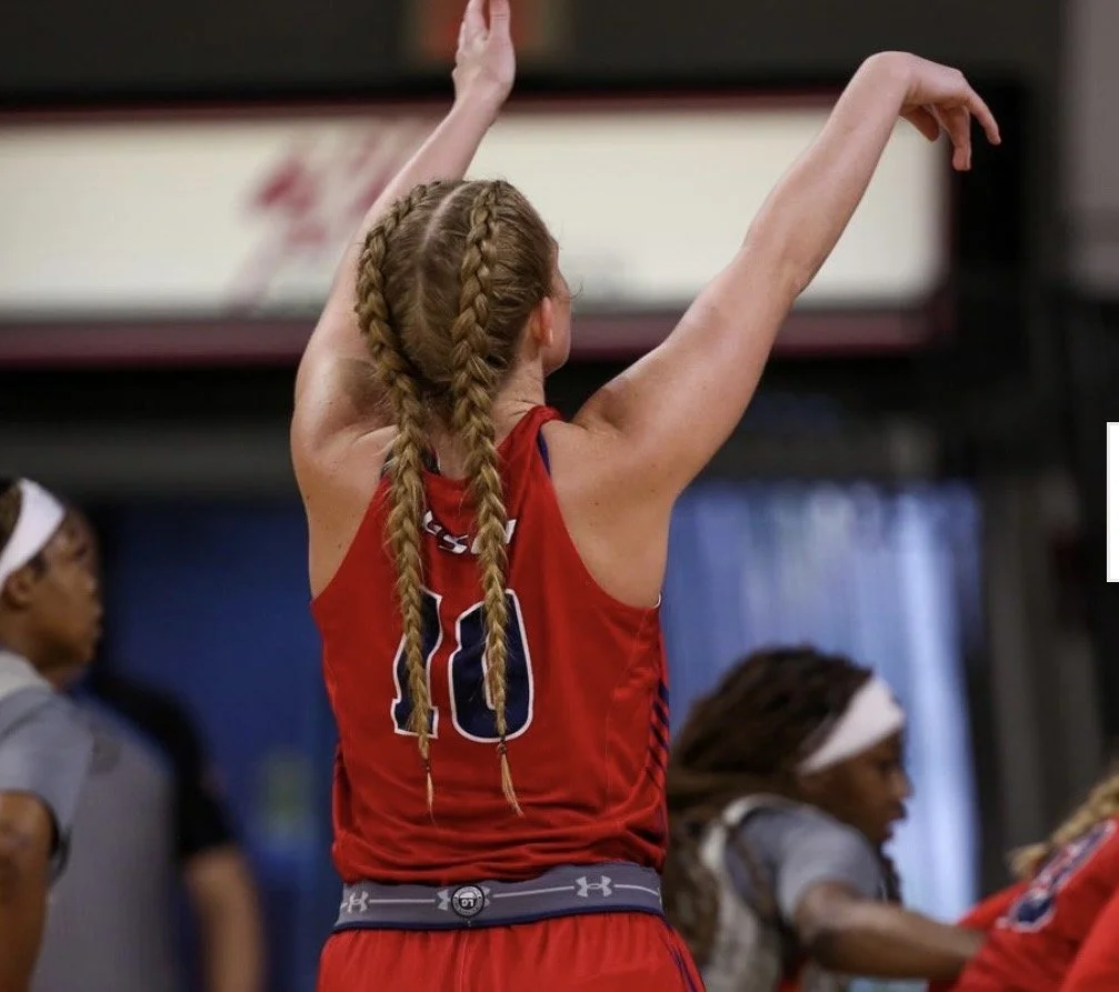 A female athlete with braided hair wearing a red sports uniform with the number 10, standing indoors with arms raised, possibly in a volleyball or basketball game, with other players and a coach in the background.