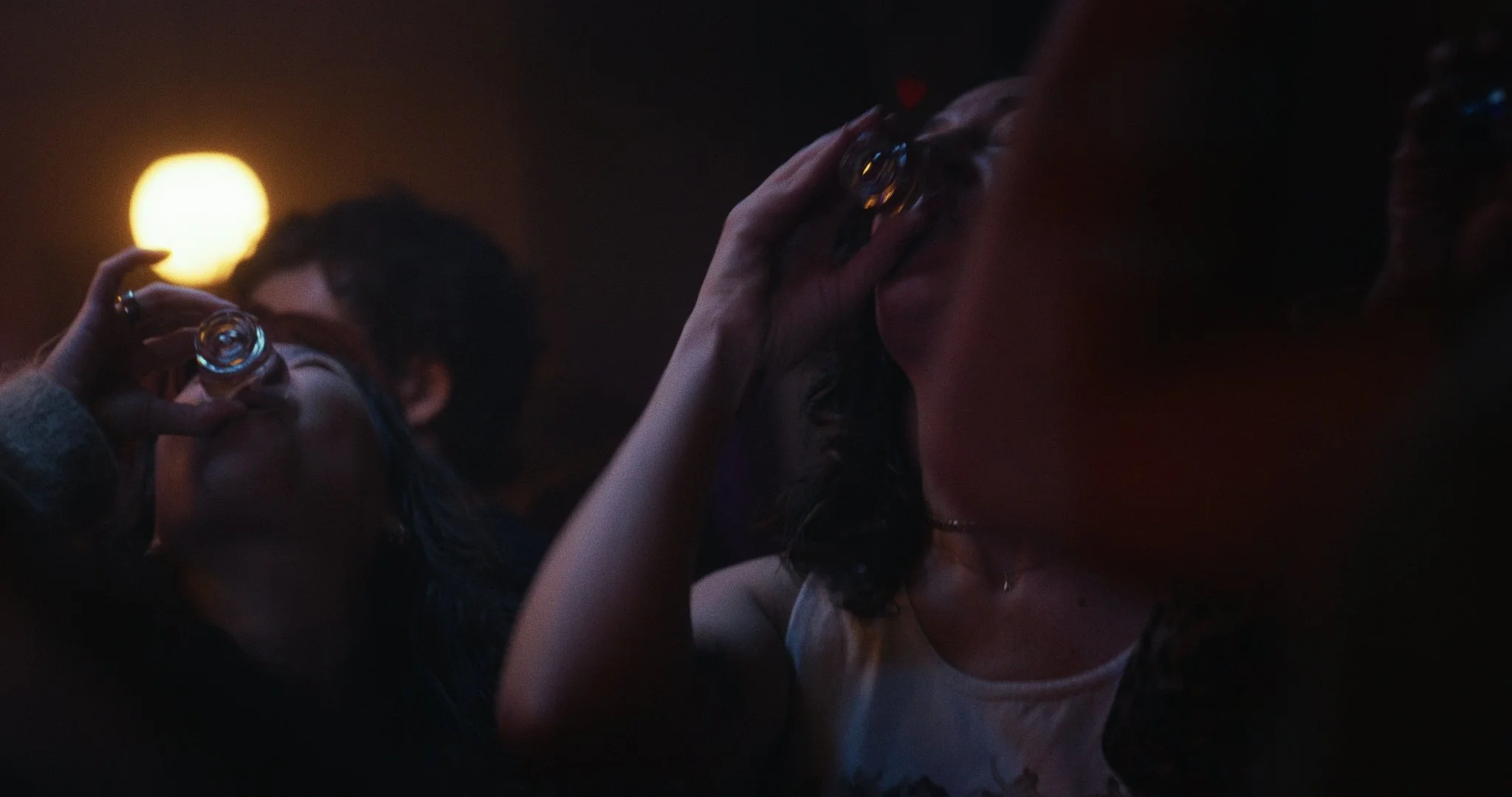 Two women laughing and drinking shots in a dark bar or club setting, with warm lighting and a round light in the background.