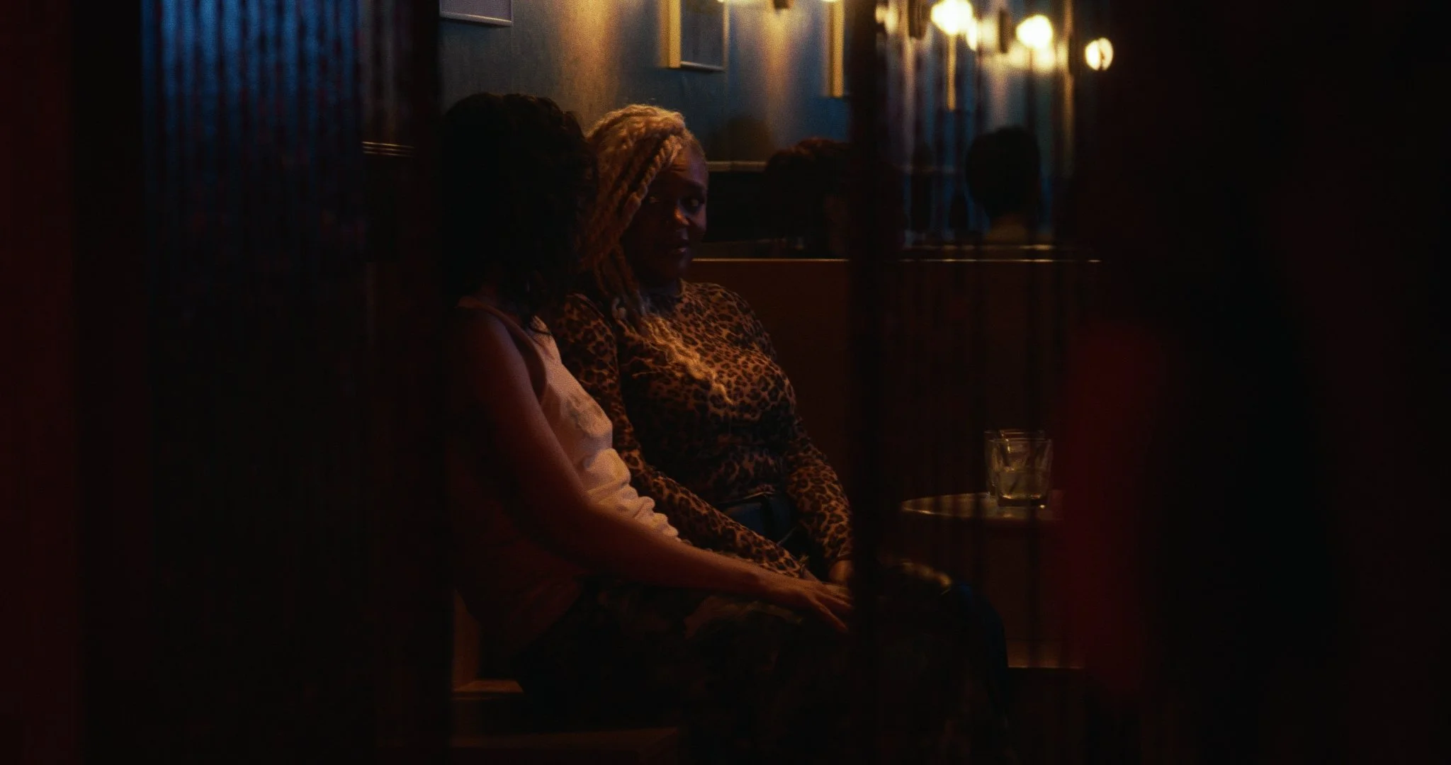 Two women sitting on a bench in a dimly lit room, one with blonde dreadlocks wearing a leopard print top and the other with dark curly hair wearing a light-colored top.