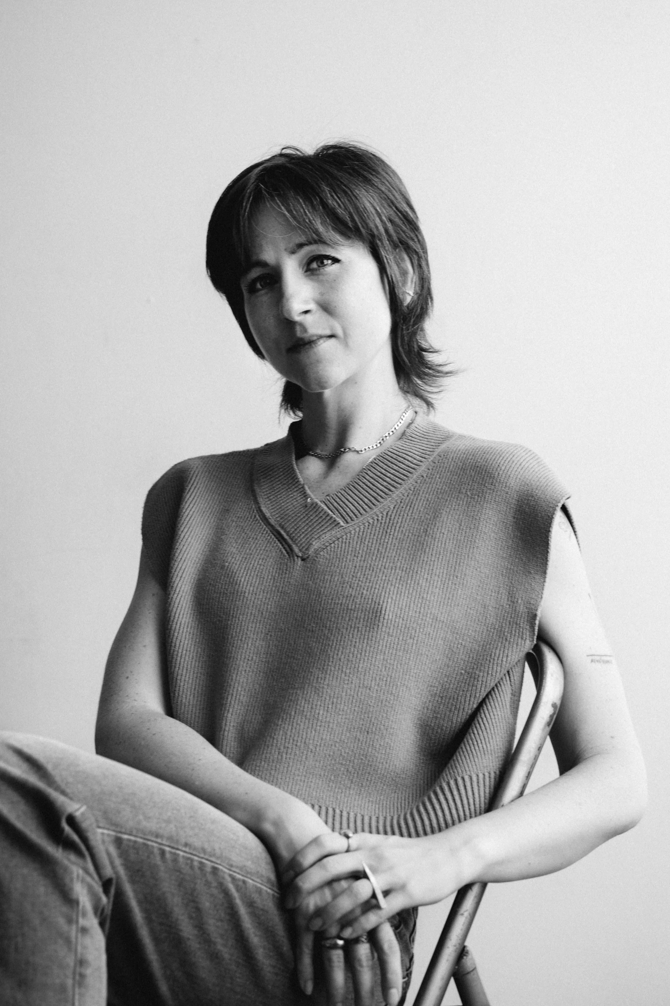 Black and white photo of a woman with short hair, wearing a sleeveless sweater, sitting on a chair with her hands clasped. She has a chain necklace and a small tattoo on her left arm.