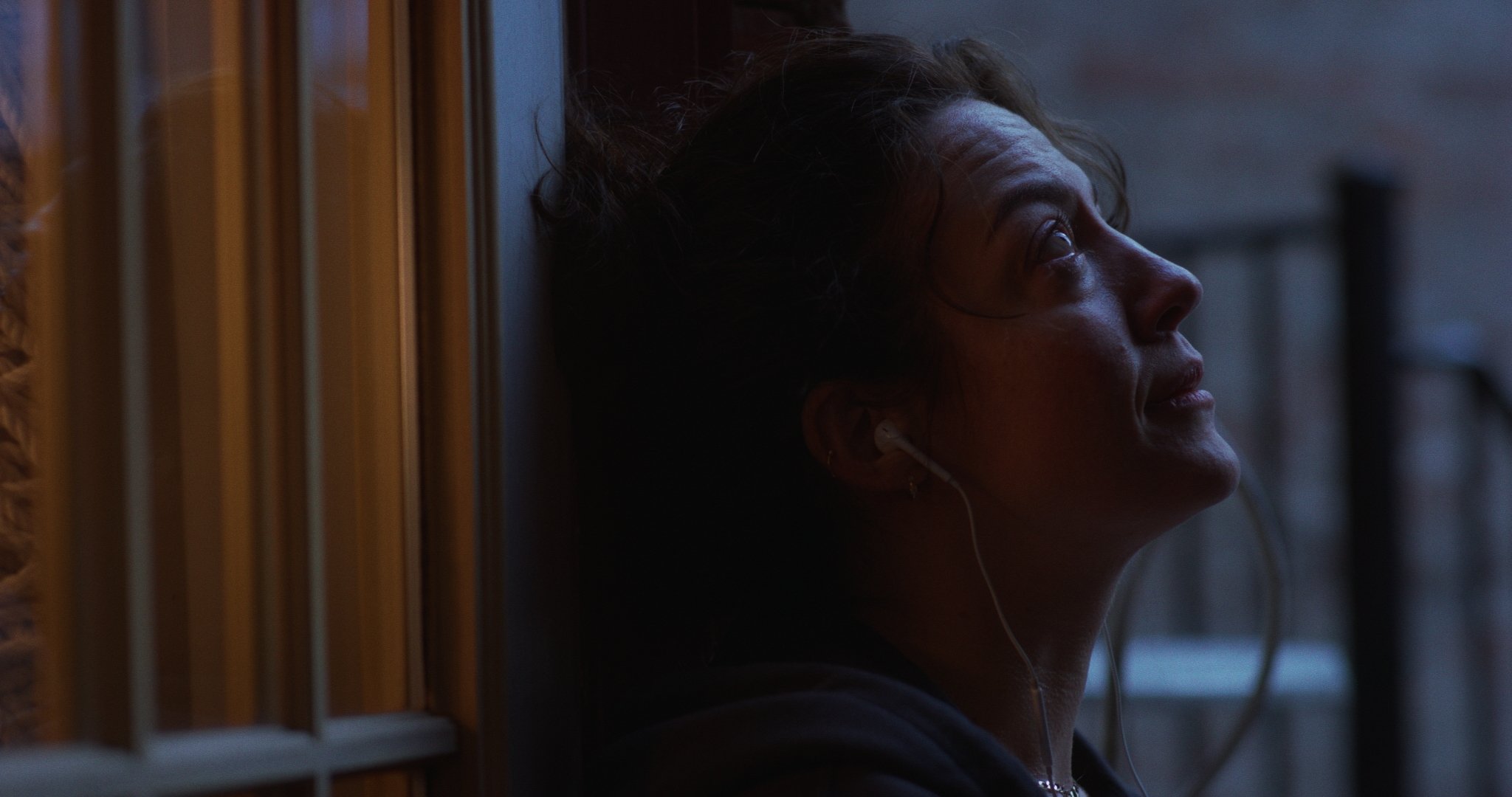 A woman with earbuds leaning against a wall, looking upward with a peaceful expression.