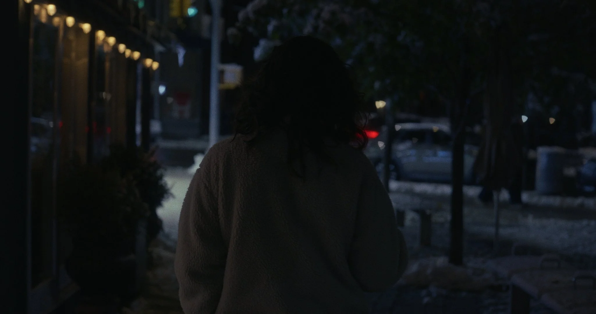 A person with dark, curly hair standing outside on a dimly lit street at night, wearing a cozy coat.