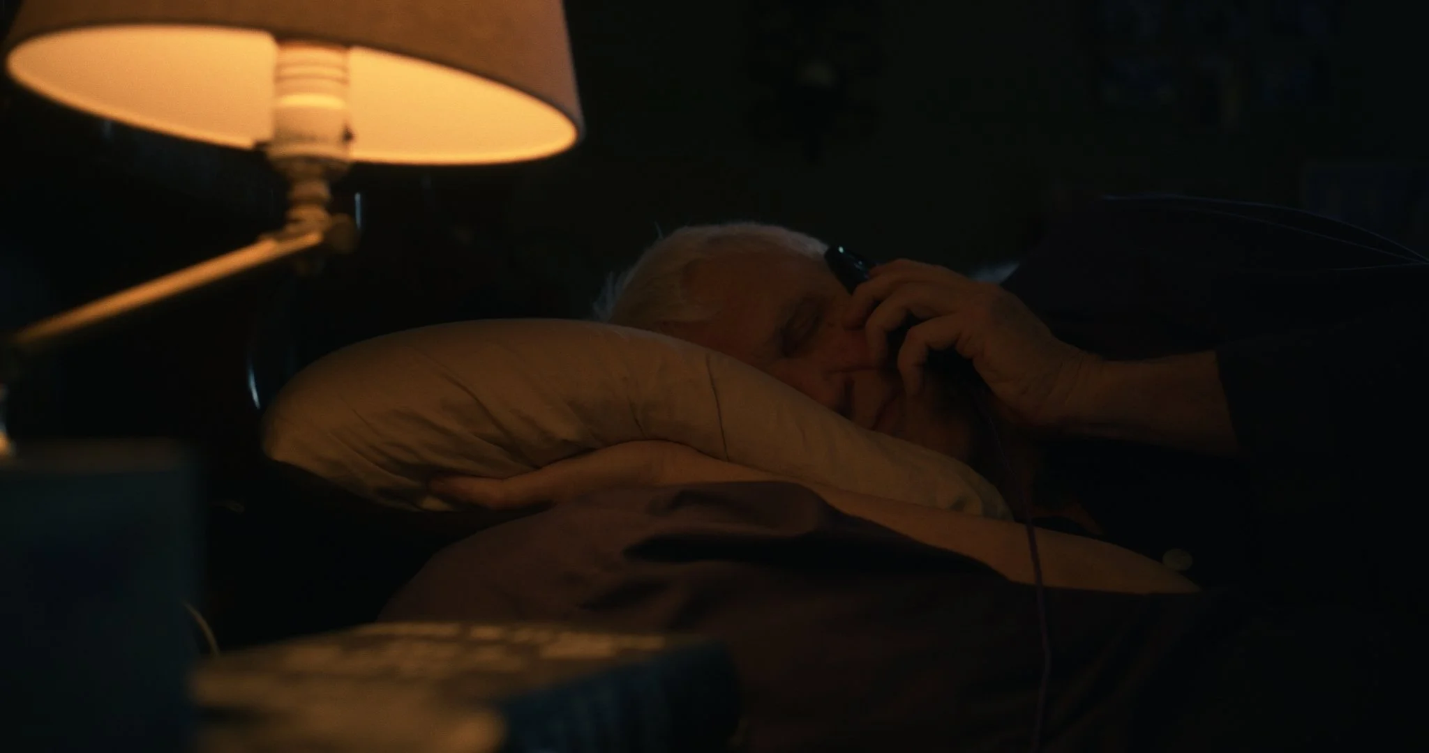An elderly man sleeping in a bed at night, illuminated by a bedside lamp, with his hand resting near his face, using a mobile phone.