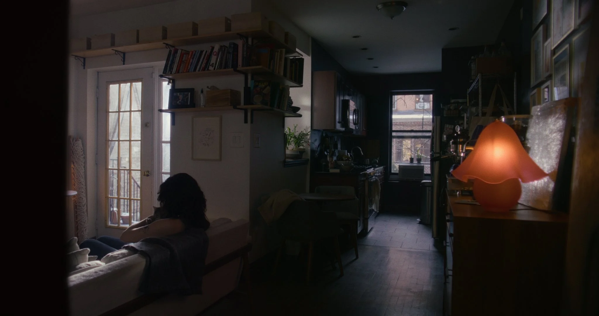 A dimly lit apartment interior with a person sitting on a bed near the door, a kitchen with dark cabinetry, a window, and a glowing orange lamp on a side table.