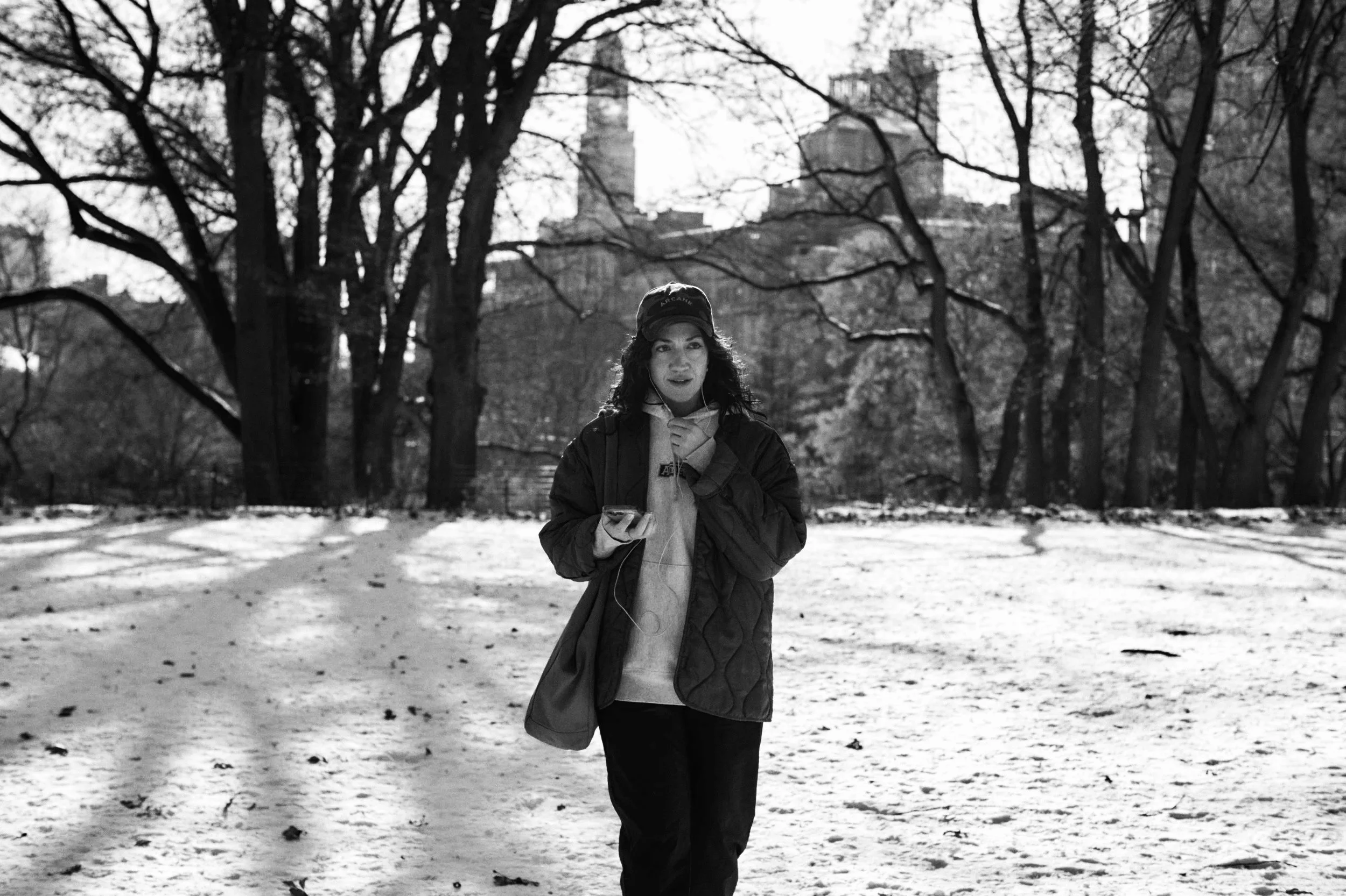 A woman walking in a snow-covered park during daytime, holding a phone and wearing a cap, jacket, and hoodie.