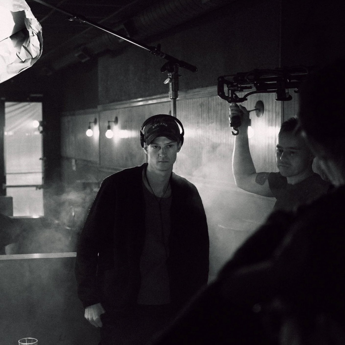 A young man wearing headphones and a cap standing in front of a camera crew, with one crew member holding a boom microphone and another operating a camera, in a dimly lit indoor setting.