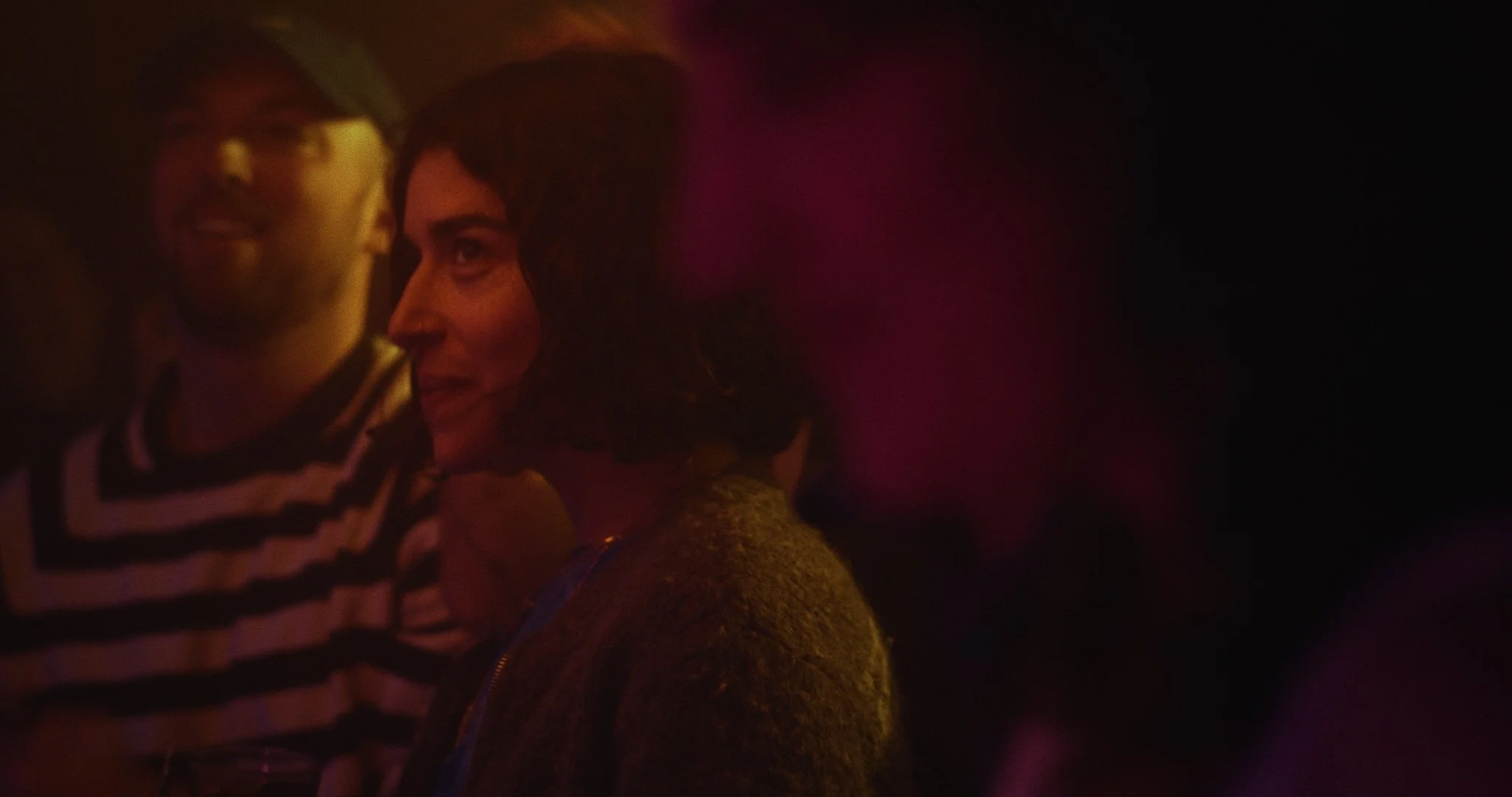 A woman with dark hair and a patterned top engaging in conversation in a dimly lit setting.