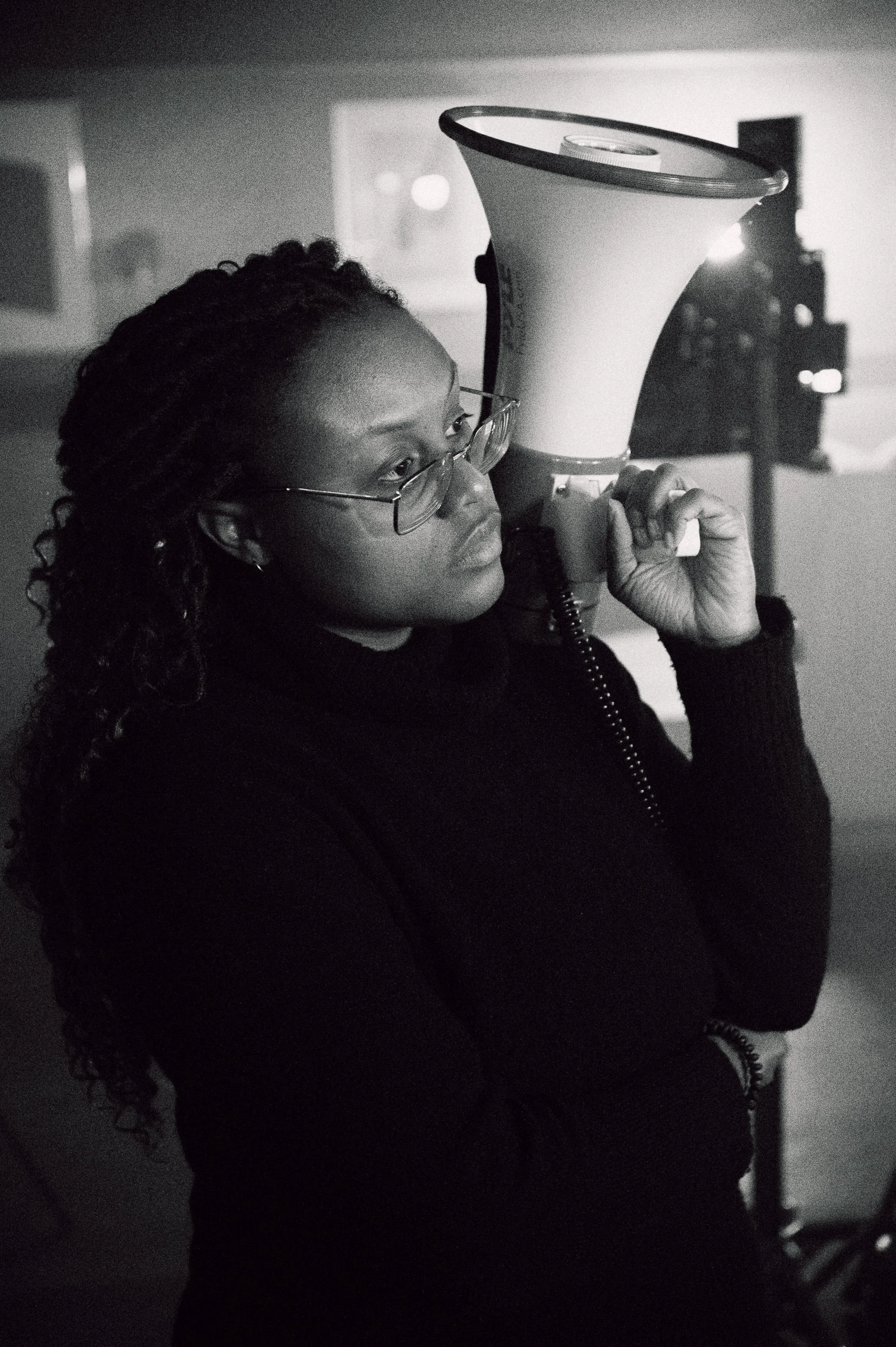 A Black woman with curly hair, wearing glasses and a black sweater, holding a megaphone on her shoulder indoors.
