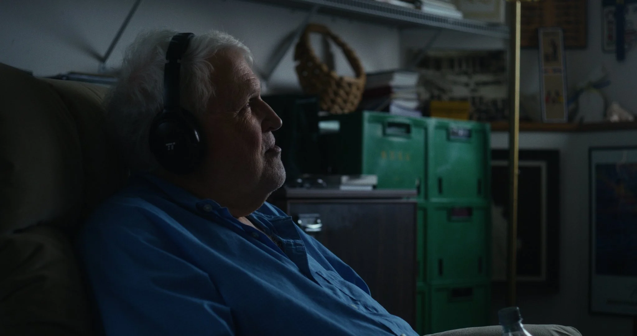 An elderly man with white hair, wearing a blue shirt, sitting on a couch and listening to music with large black headphones in a dimly lit room.