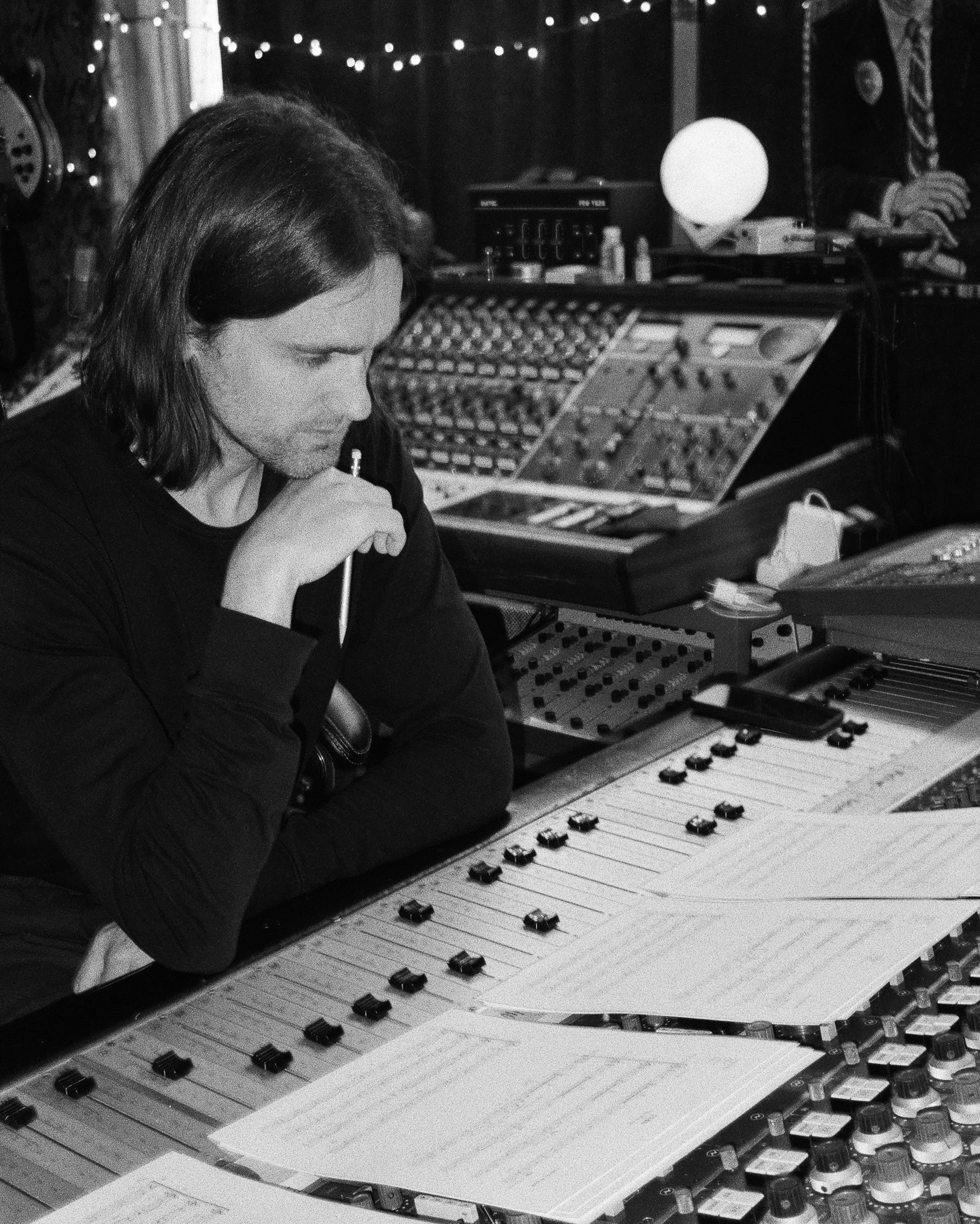 A man with shoulder-length hair sits at a mixing console in a recording studio, surrounded by audio equipment, with sheet music in front of him and a contemplative expression.