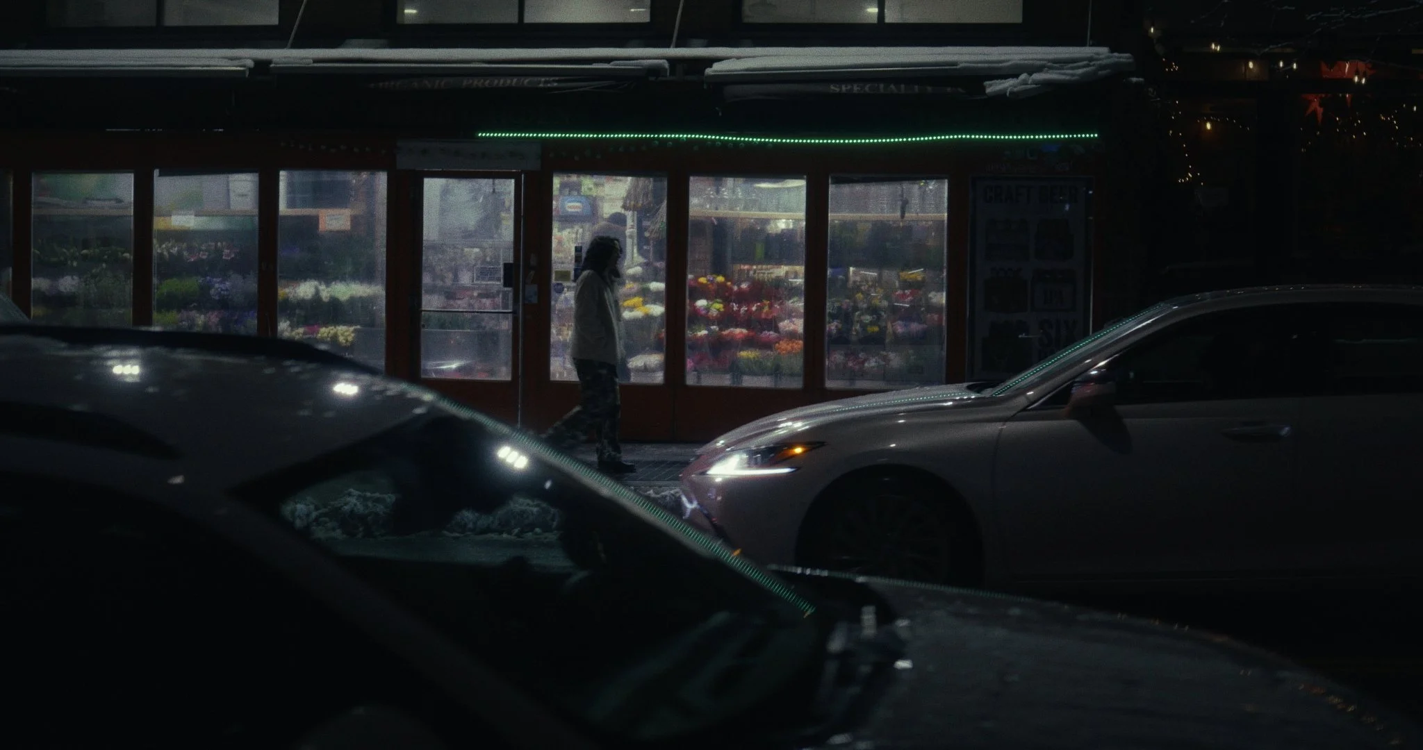 Person walking past a flower shop at night, with cars parked in front.