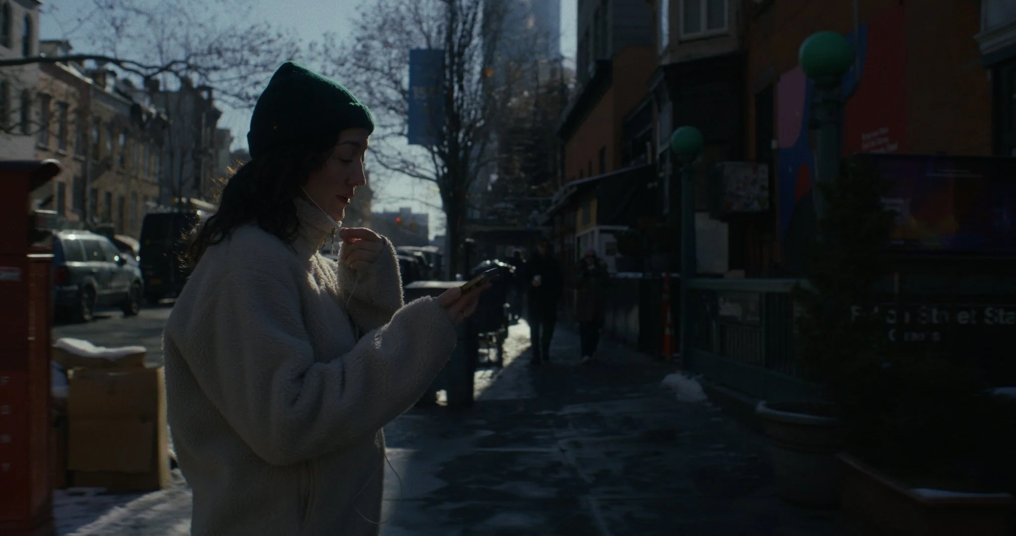 A woman wearing a dark green beanie and beige sweater standing on a city sidewalk, looking at her phone and listening to earphones, with a background of parked cars, leafless trees, and buildings.