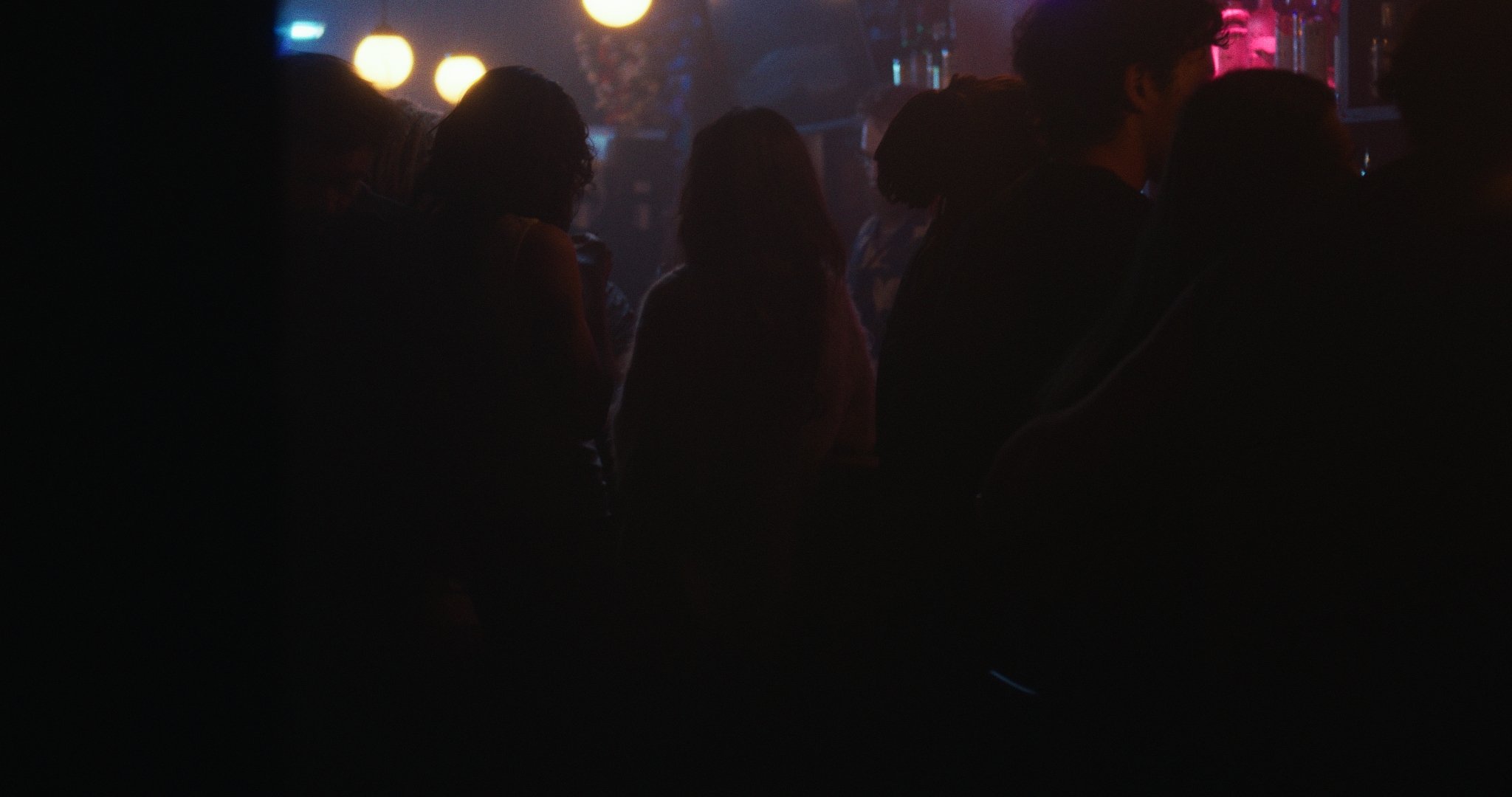 Silhouettes of people sitting and standing in a dimly lit bar or nightclub with colorful lights and neon signs in the background.