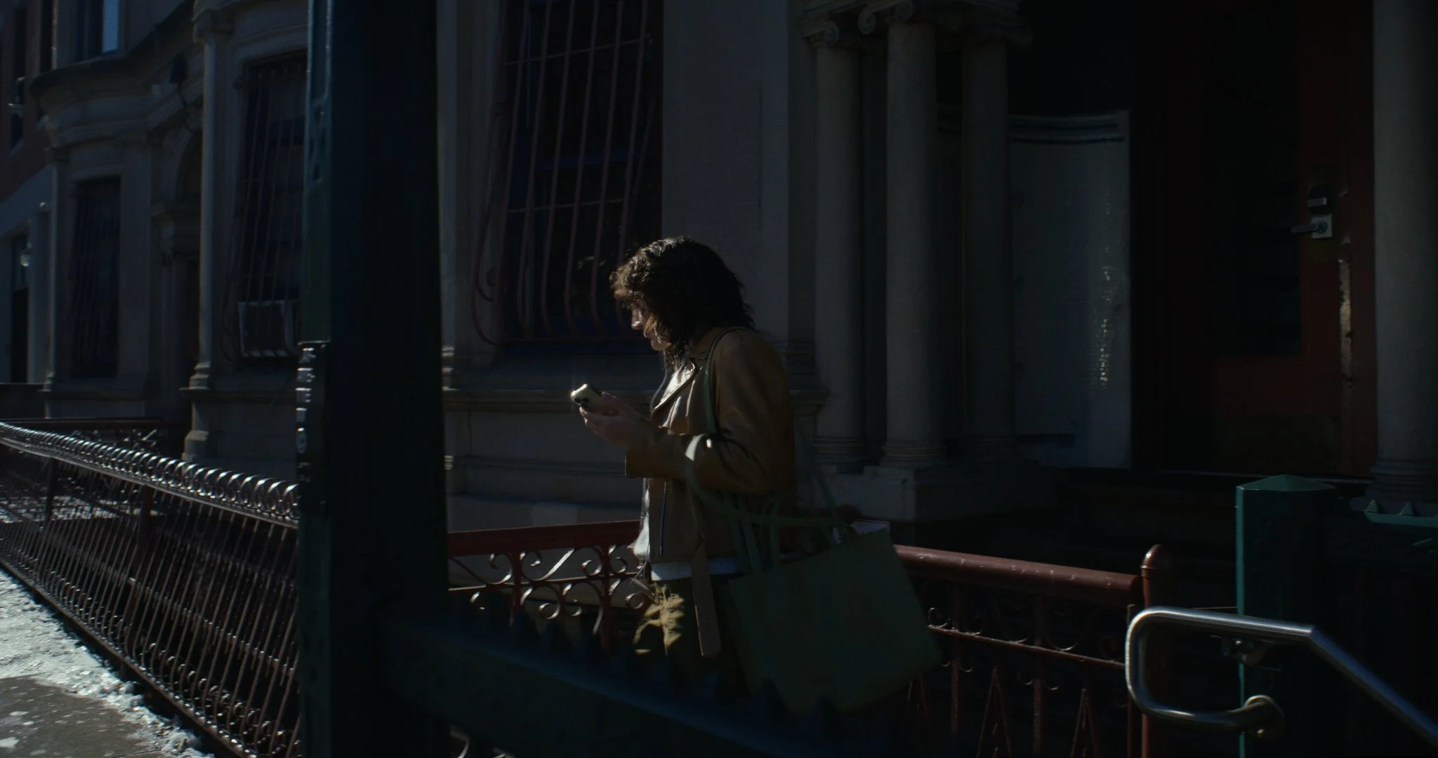 A woman with dark, curly hair wearing a beige leather jacket and brown pants stands outside a building, looking down at her smartphone with sunlight illuminating her face. She carries a large tote bag over her shoulder and stands near a black iron fe