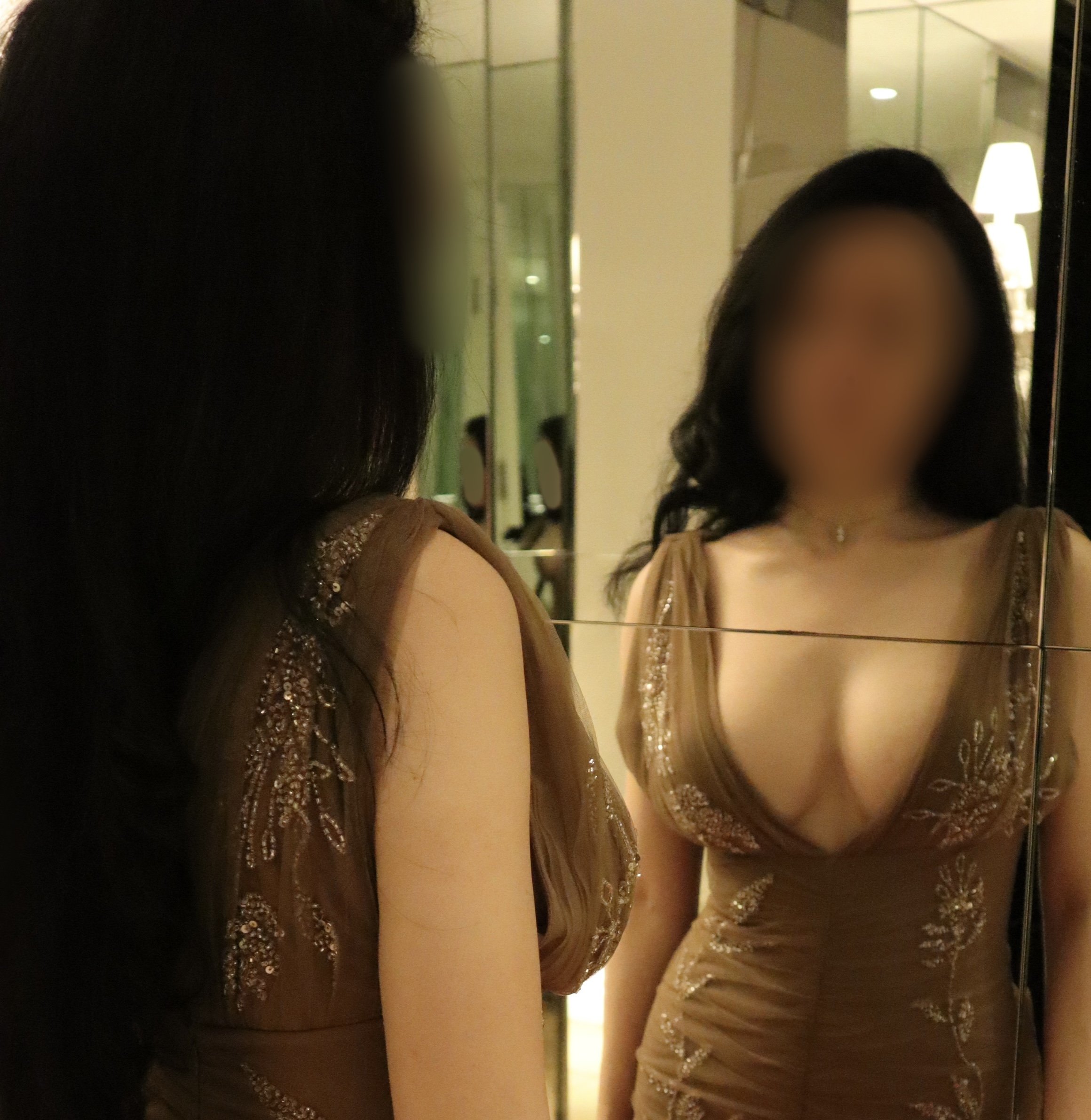 A mirror reflection of two women at an indoor event, dressed in elegant, embellished dresses, with a blurred background of warm lighting and modern interior decor.