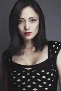 Portrait of a woman with black hair and red lipstick wearing a black top with cutout pattern against a gray background.