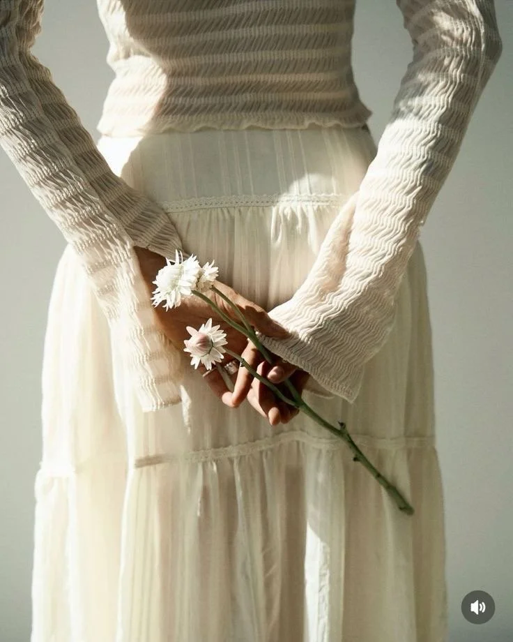 Person in a cream-colored, long-sleeved, textured top and matching skirt holding white daisies behind their back.