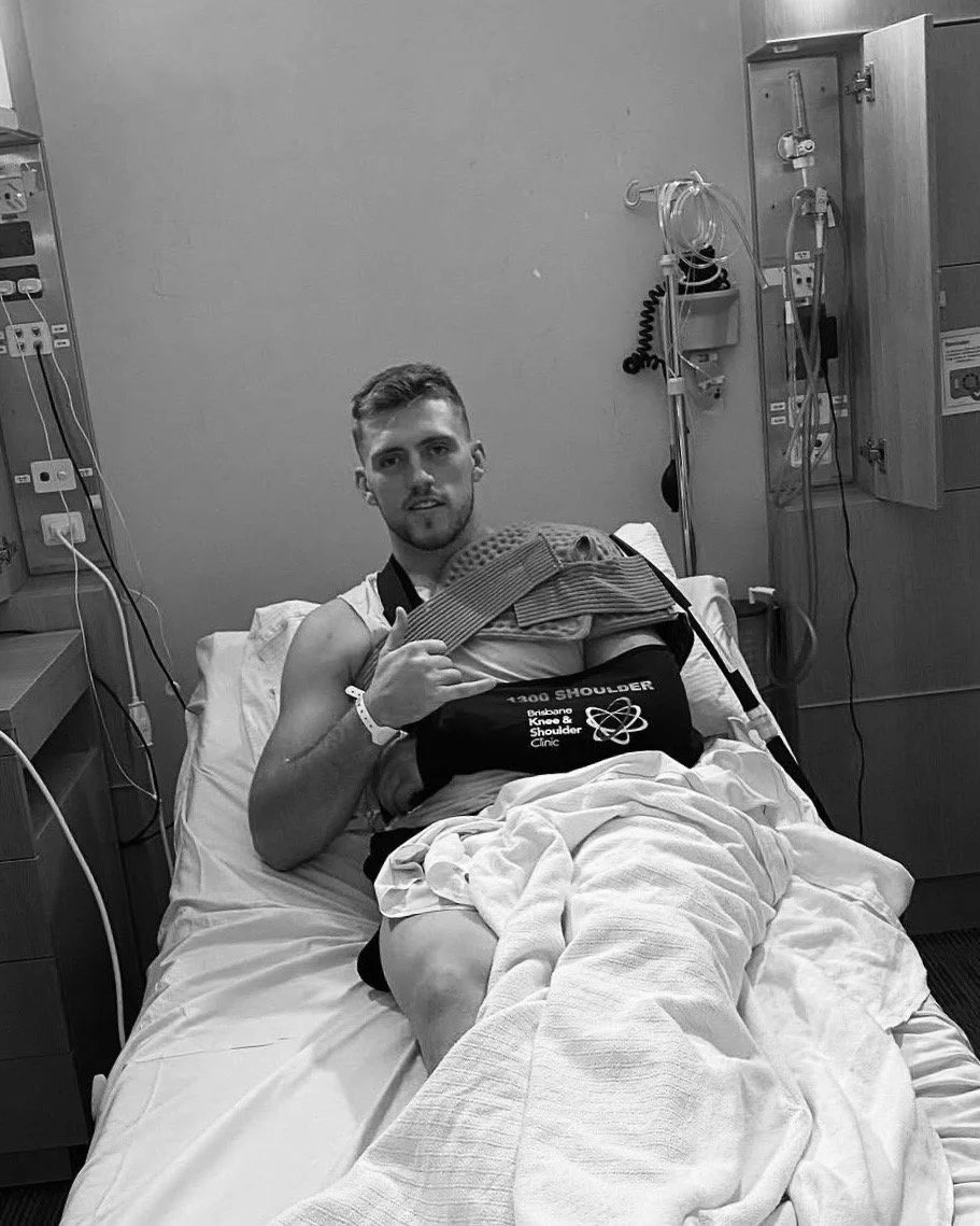 A young man in a hospital bed, giving a thumbs-up. He has a shoulder sling on his left arm, a hospital bracelet on his right wrist, and medical equipment around him in a hospital room.