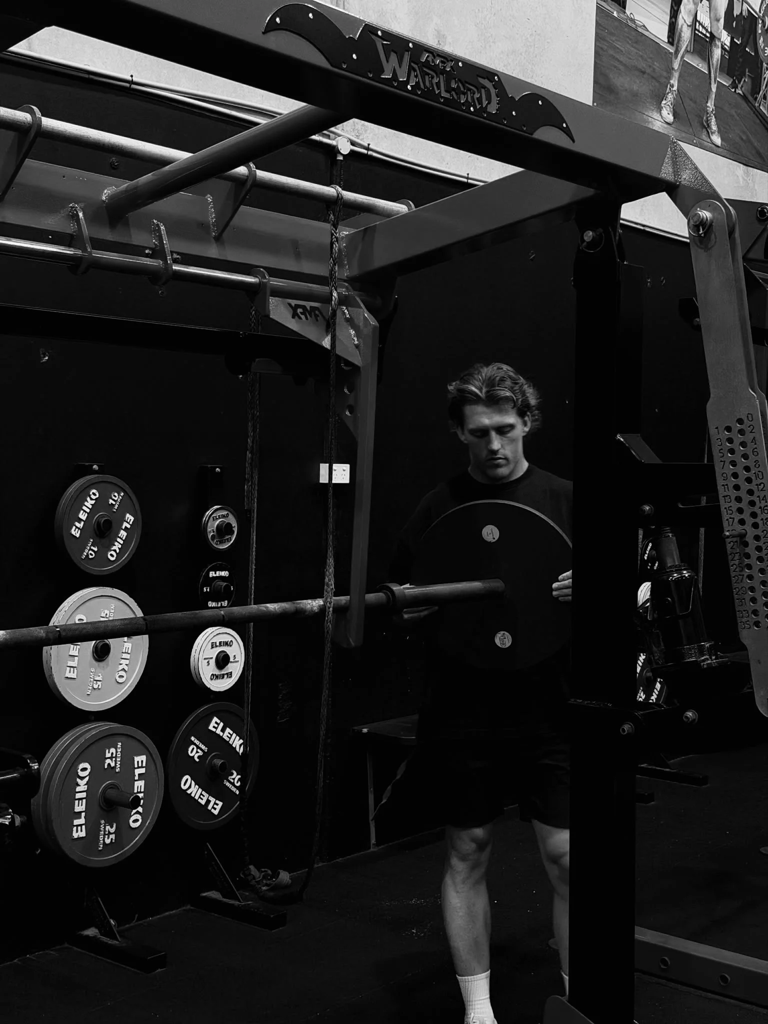 A man in a gym standing next to a wall with weight plates, barbell, and gym equipment. He is wearing a black t-shirt, shorts, and white socks.