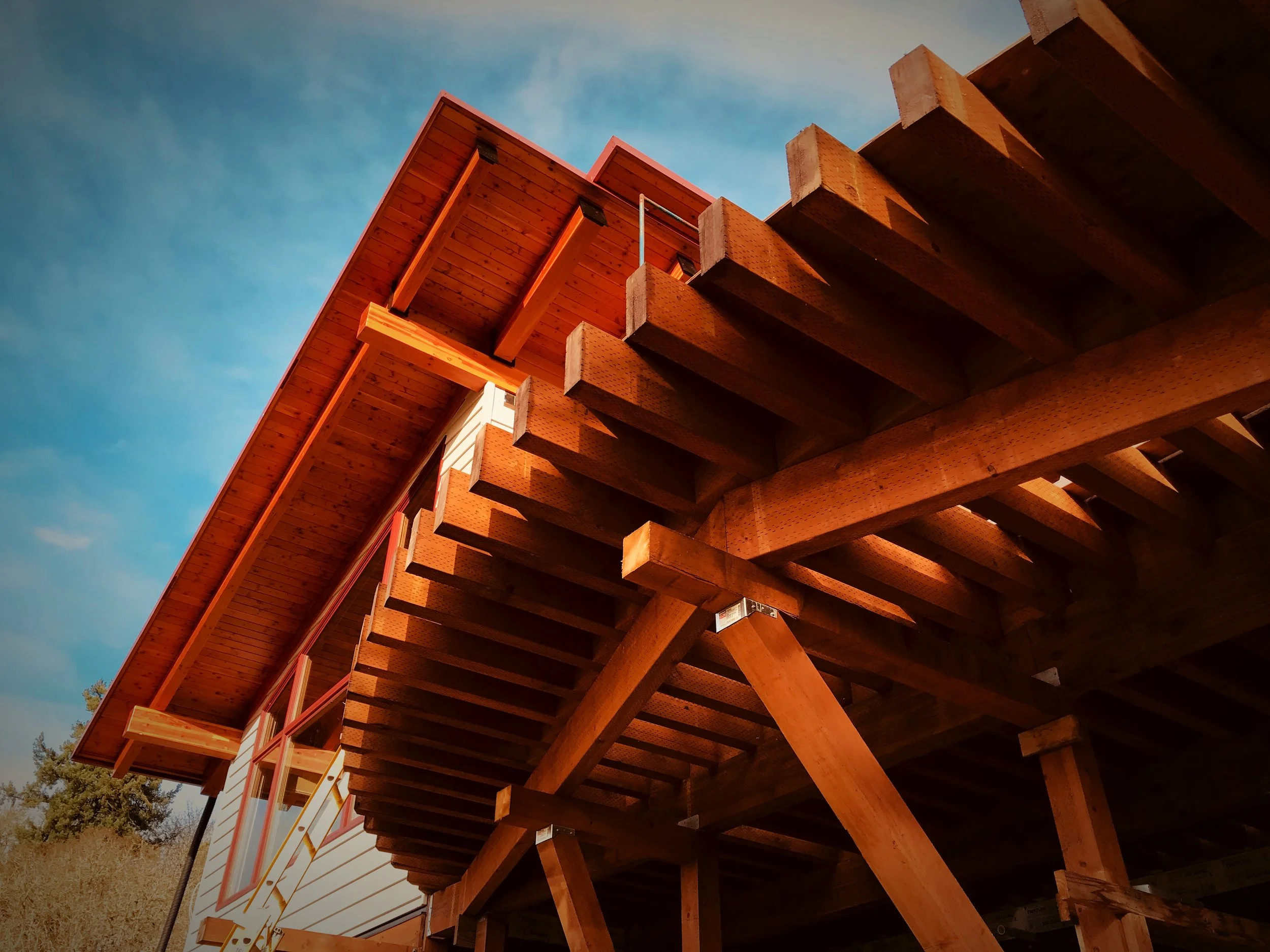 A low-angle view of a modern house with large wooden beams supporting an overhanging roof, set against a blue sky with some wispy clouds. The house has large windows and white exterior walls.