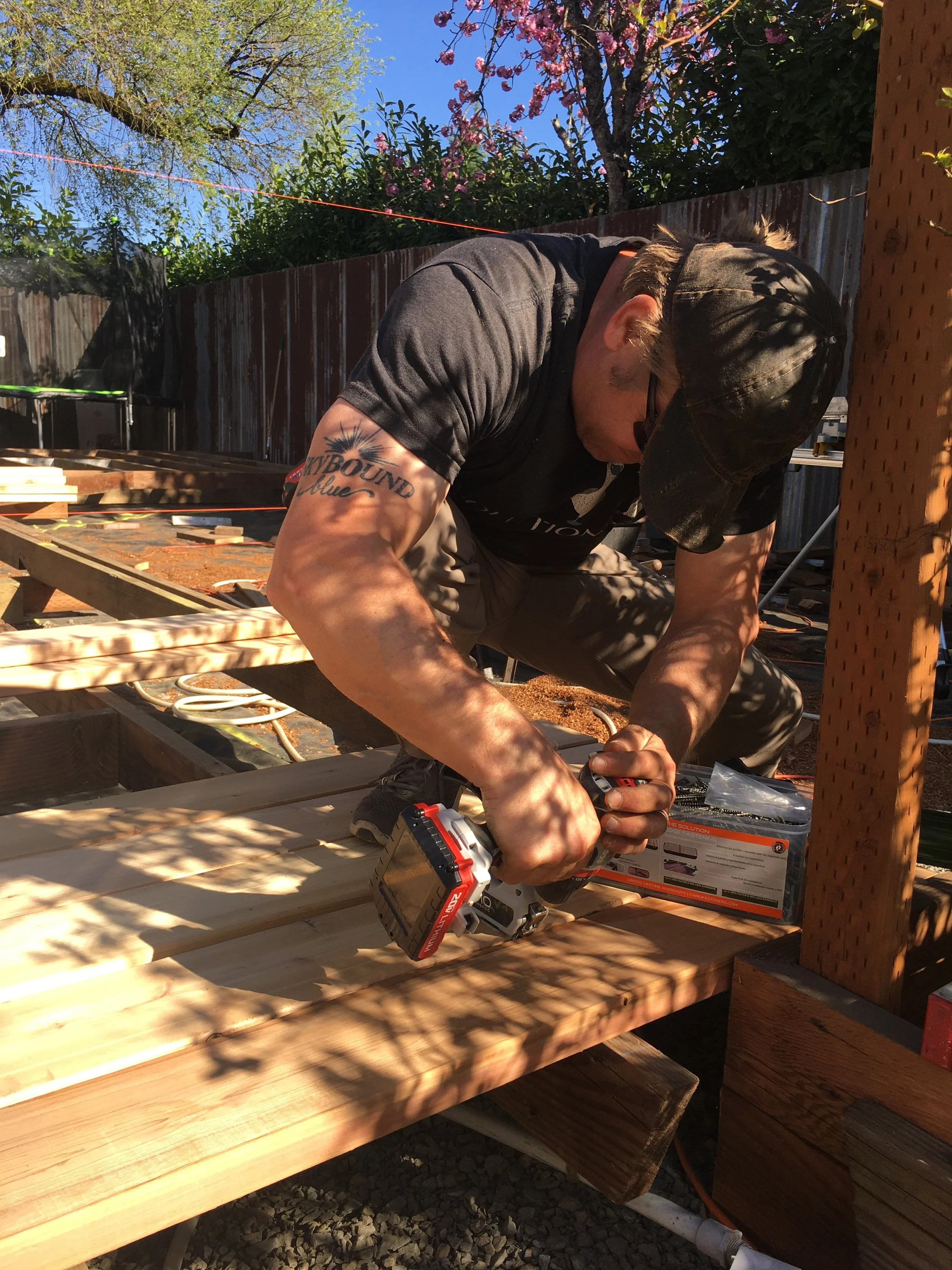 Ark Built Carpentry founder Matt Behnke wearing a cap, sunglasses, and a black t-shirt is kneeling on a wooden deck, working with a cordless drill on a large custom designed deck.